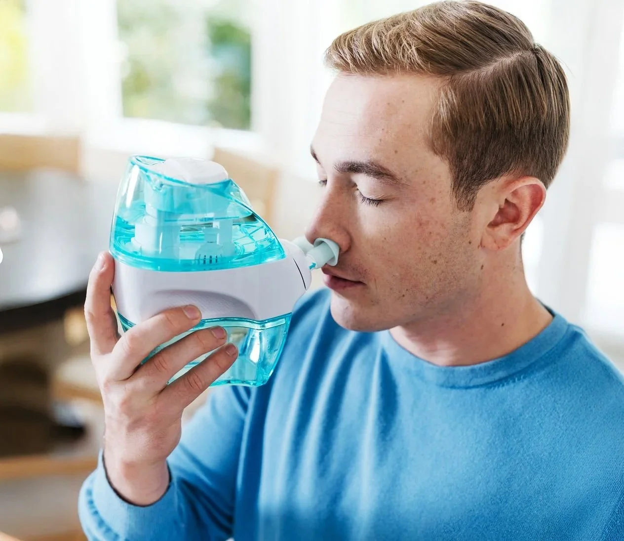 A young man with short light brown hair and a blue shirt uses a nebulizer machine to breathe medication, with his eyes closed.