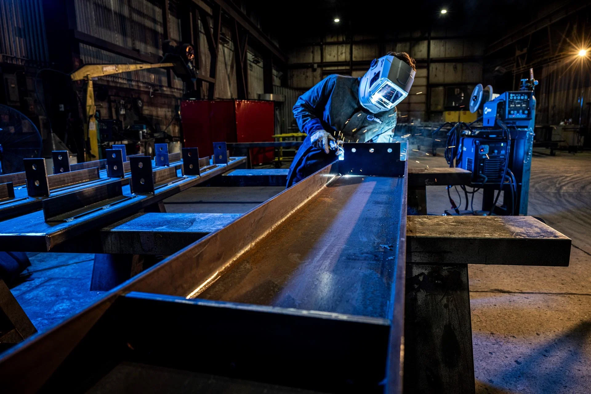 A welder wearing a protective helmet and gloves working on a metal frame inside a factory or workshop. Sparks are flying from the welding process, with welding equipment nearby and a wooden workspace.