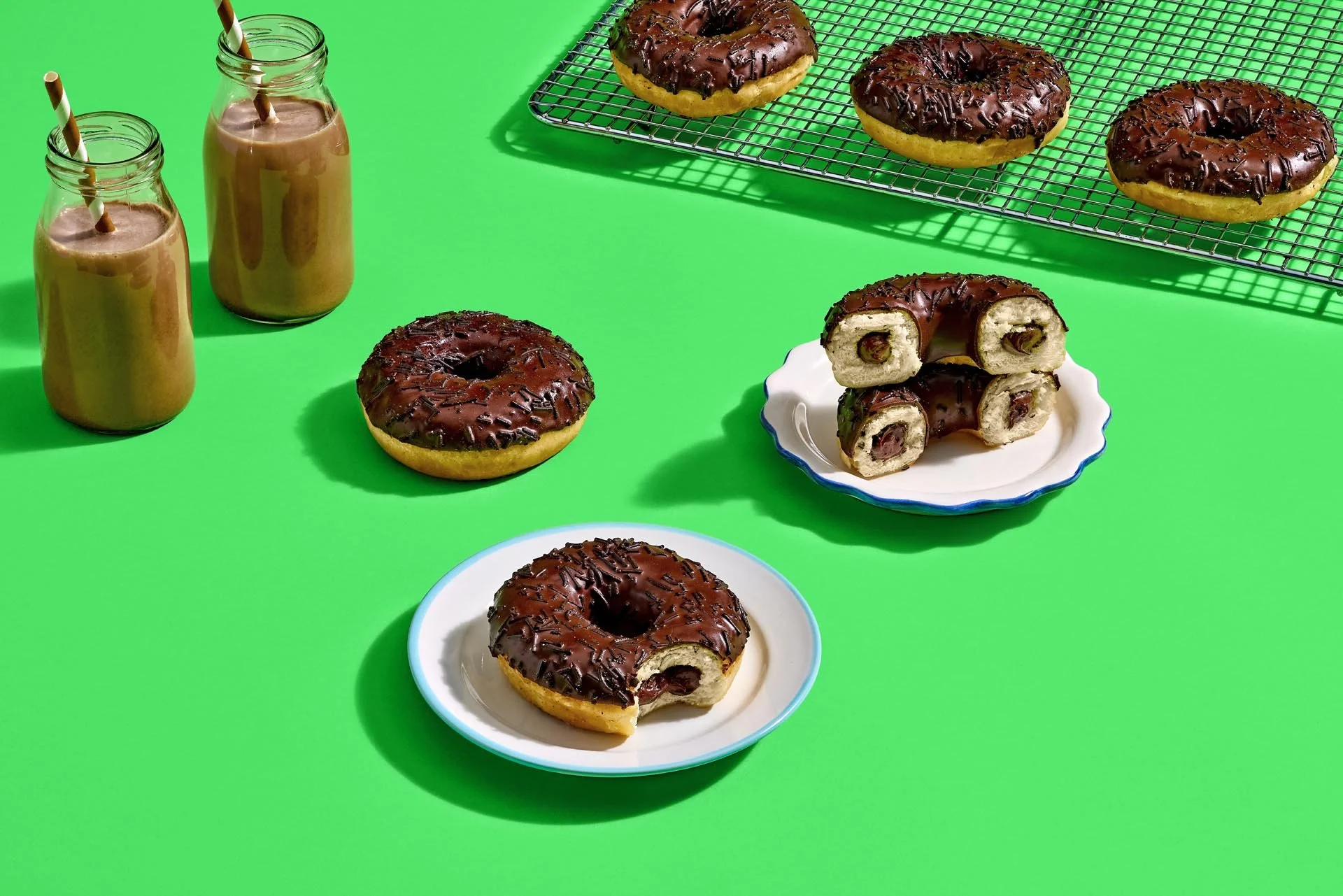 Chocolate-glazed donuts with sprinkles on a wire rack and plates with donut halves on a bright green surface, accompanied by two bottles of chocolate milk with striped straws.