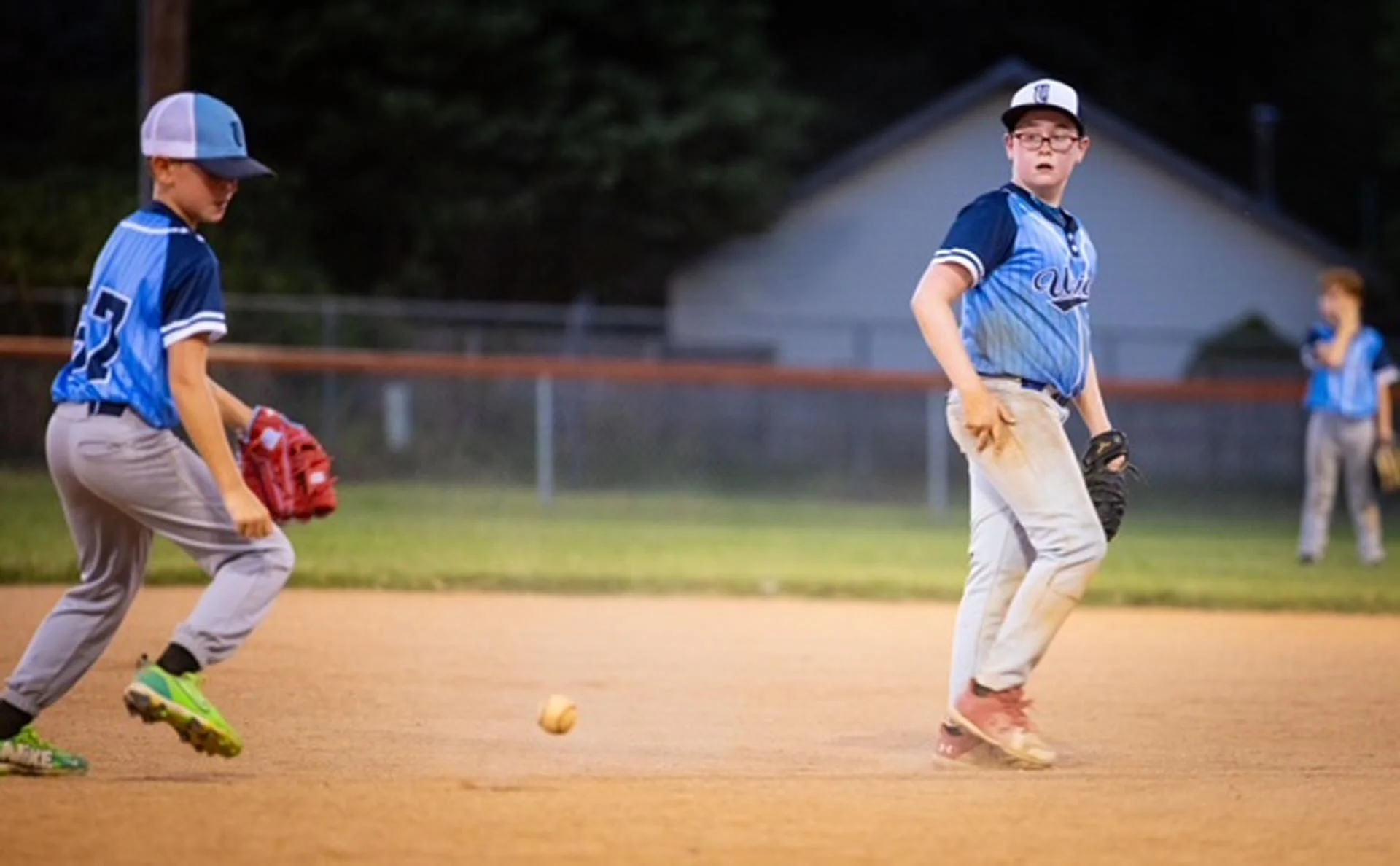 Youth baseball game at dusk with players on the field, one in a blue and white uniform preparing to catch, another in a blue and gray uniform, and a girl in the background near the fence.
