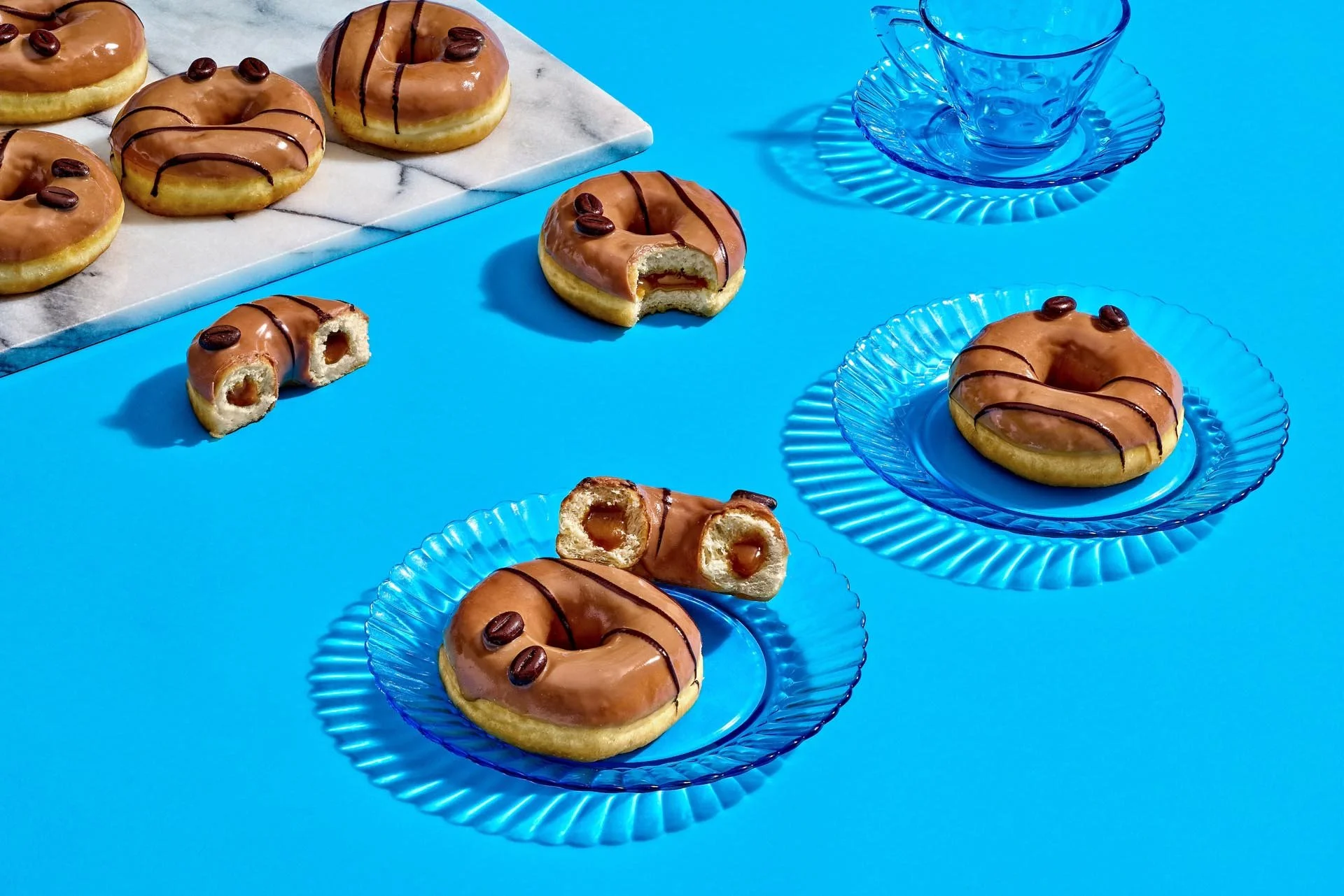 Several donuts with chocolate glaze and coffee beans on top on blue glass plates, some donuts are partially eaten, on a bright blue background with a marble tray holding more donuts.