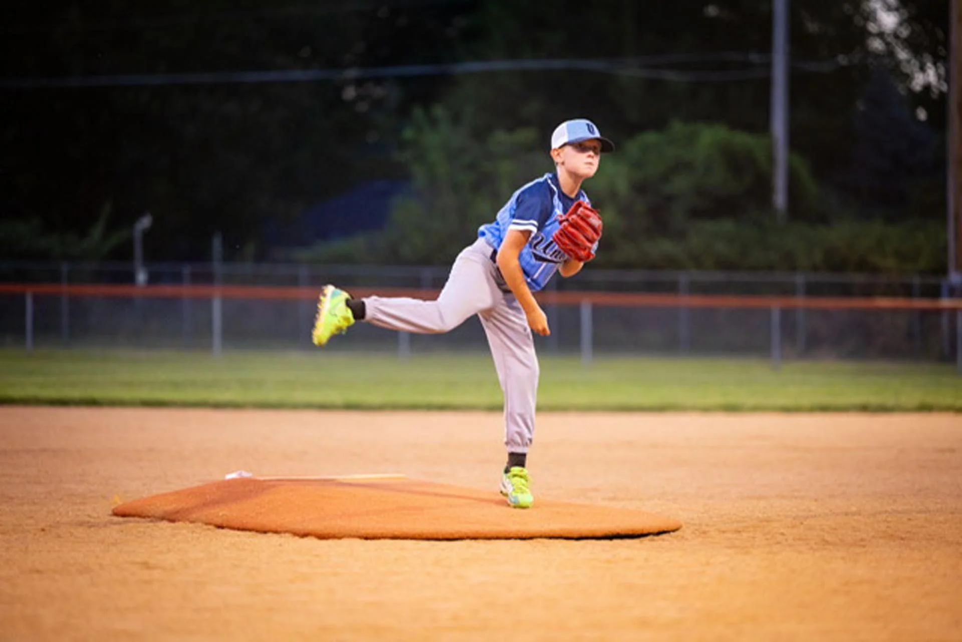 A young boy in a baseball uniform throwing a pitch on a baseball field during evening or early morning.
