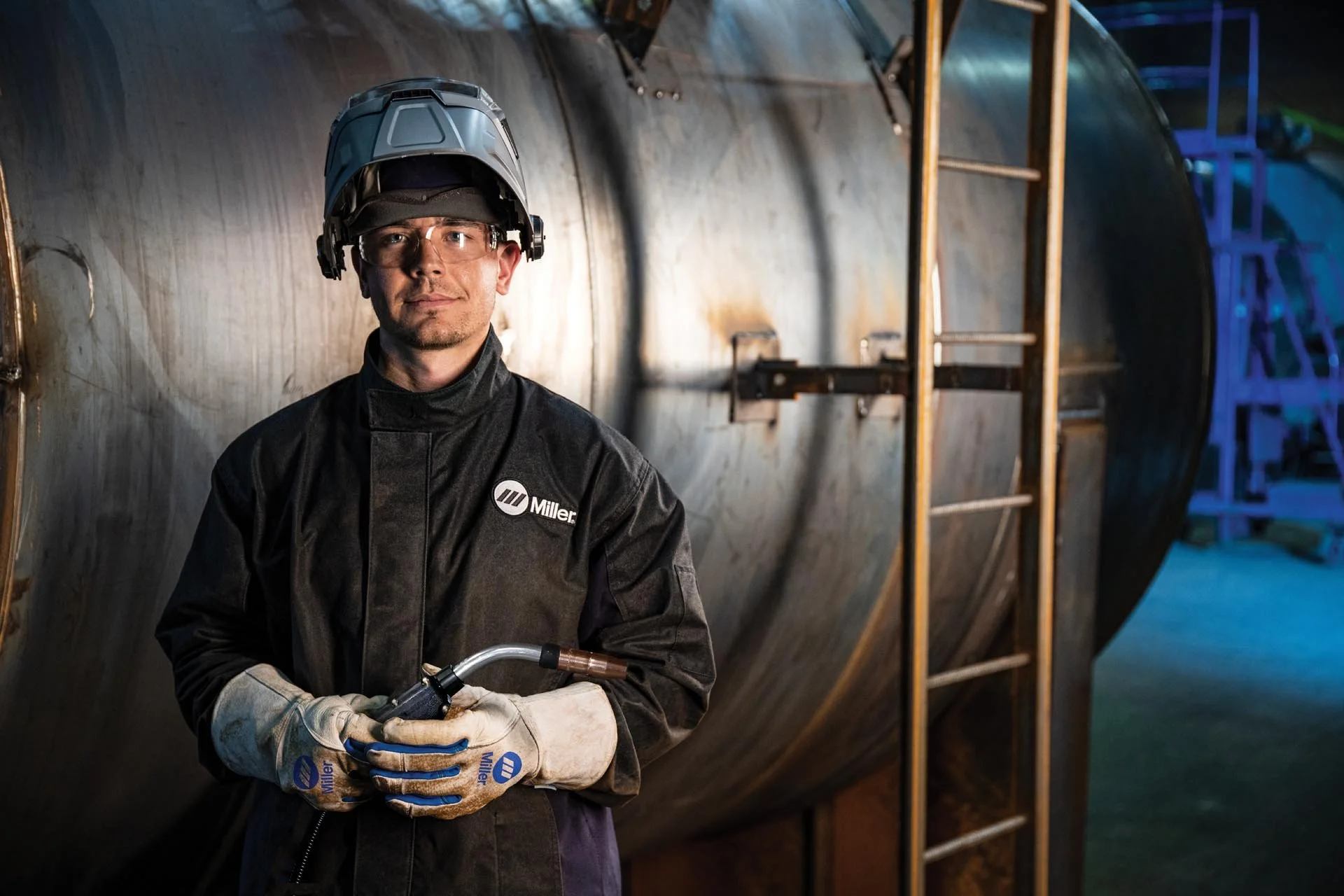 A man wearing safety glasses, a jacket with 'Miller' logo, welding gloves, and a safety helmet holding welding equipment while standing next to a large metal tank or pipe in an industrial setting.