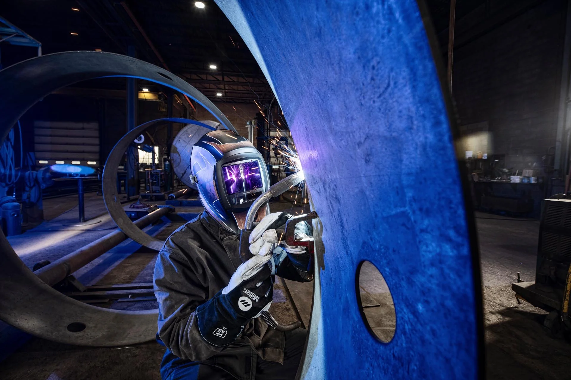 A worker wearing a welding helmet and gloves welding a large blue metal pipe, with sparks flying indoors in an industrial workshop.