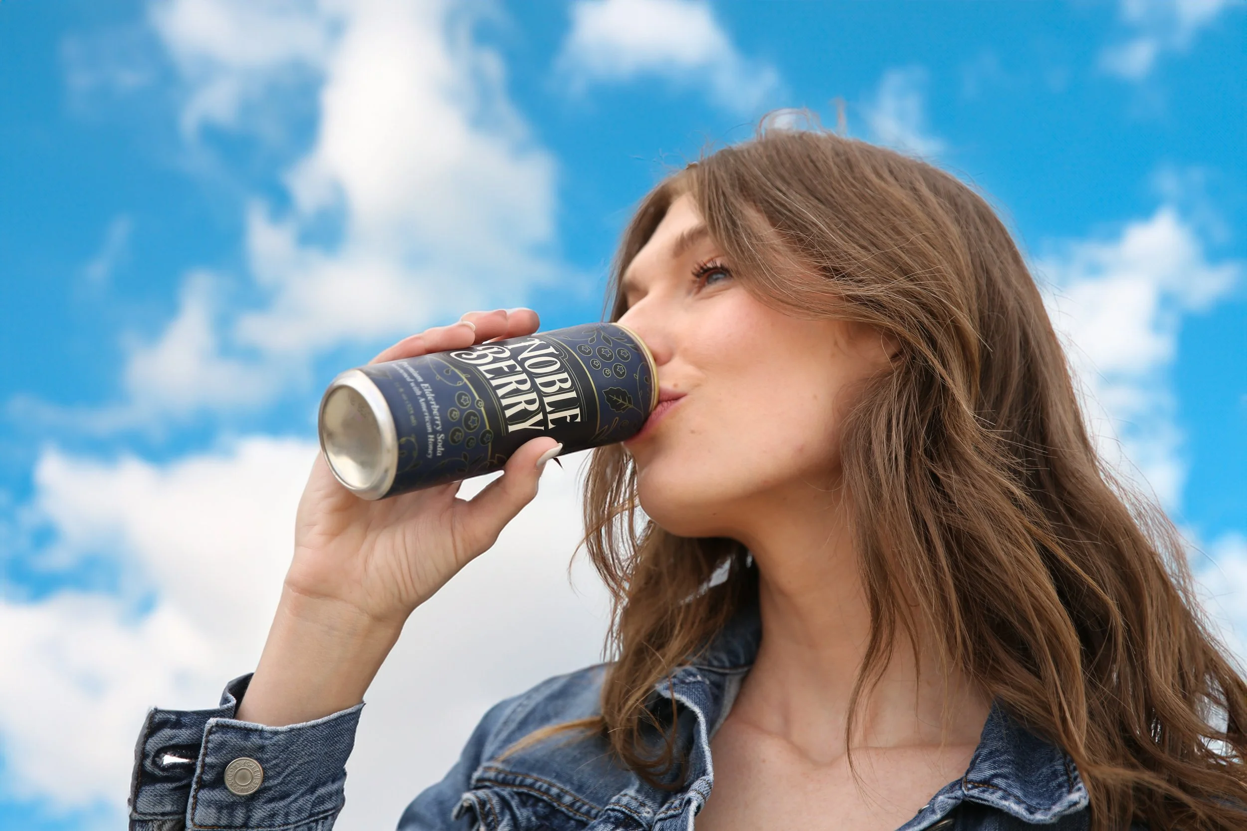 A young woman with long brown hair drinking from a can labeled 'Noble Beer' while standing outdoors under a partly cloudy sky.