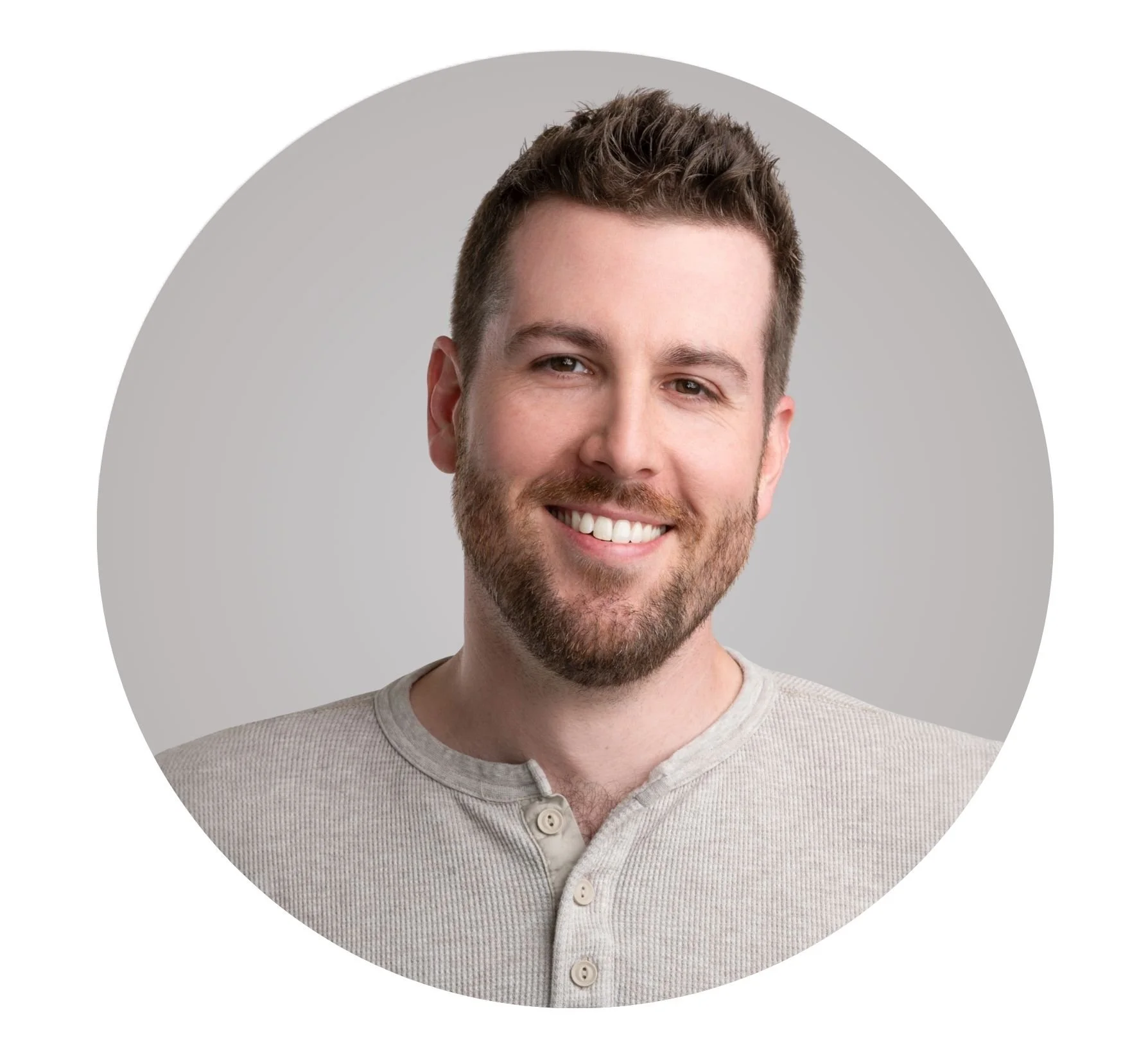 Portrait of a young man with short brown hair and a beard, smiling, wearing a light gray button-up shirt, against a gray background.