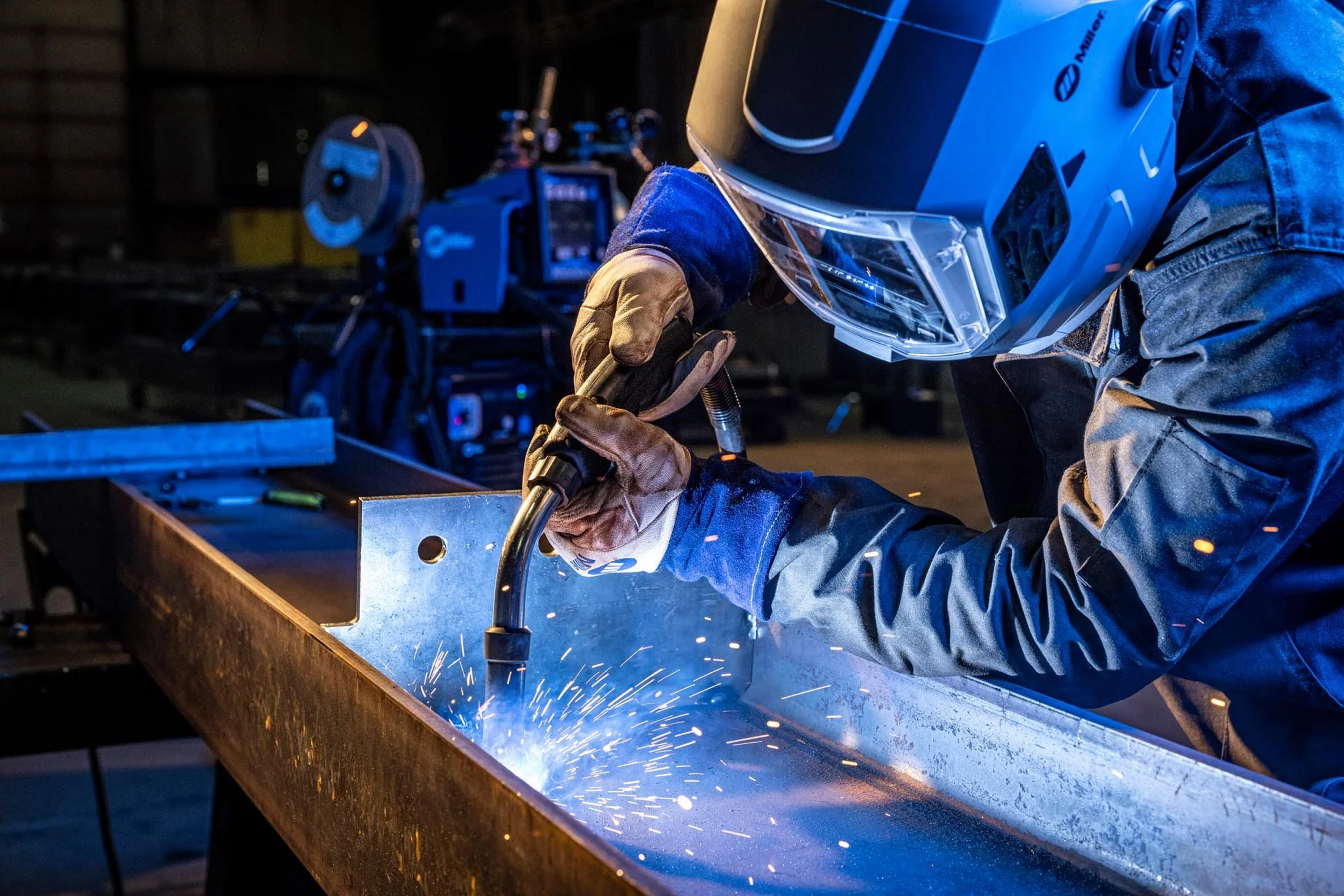 Welder welding metal on a worktable, sparks flying, in a workshop, wearing protective helmet and gloves, blue lighting.