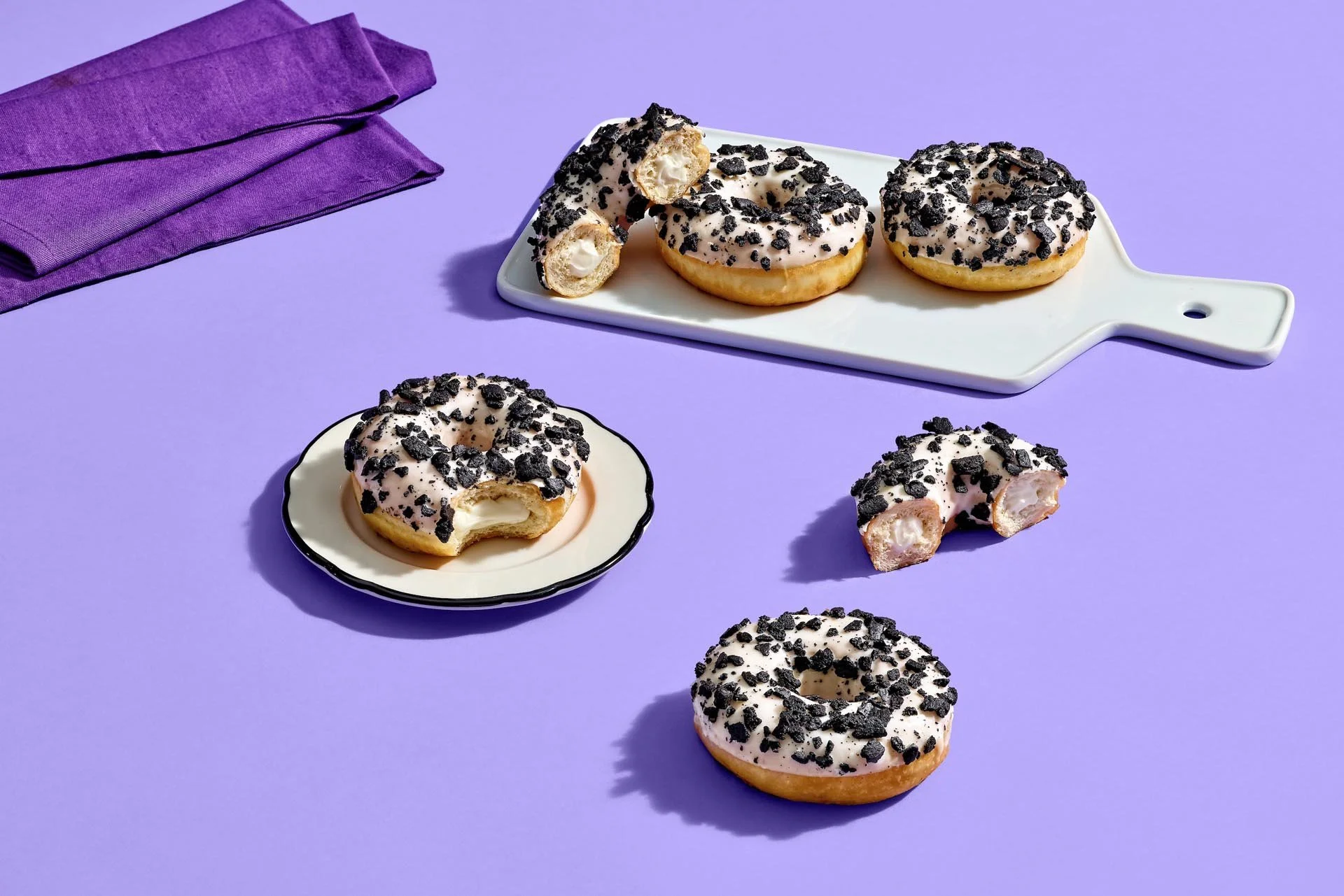 Several donuts with white icing and black cookie crumbs on top, some on small plates and others on a purple background.