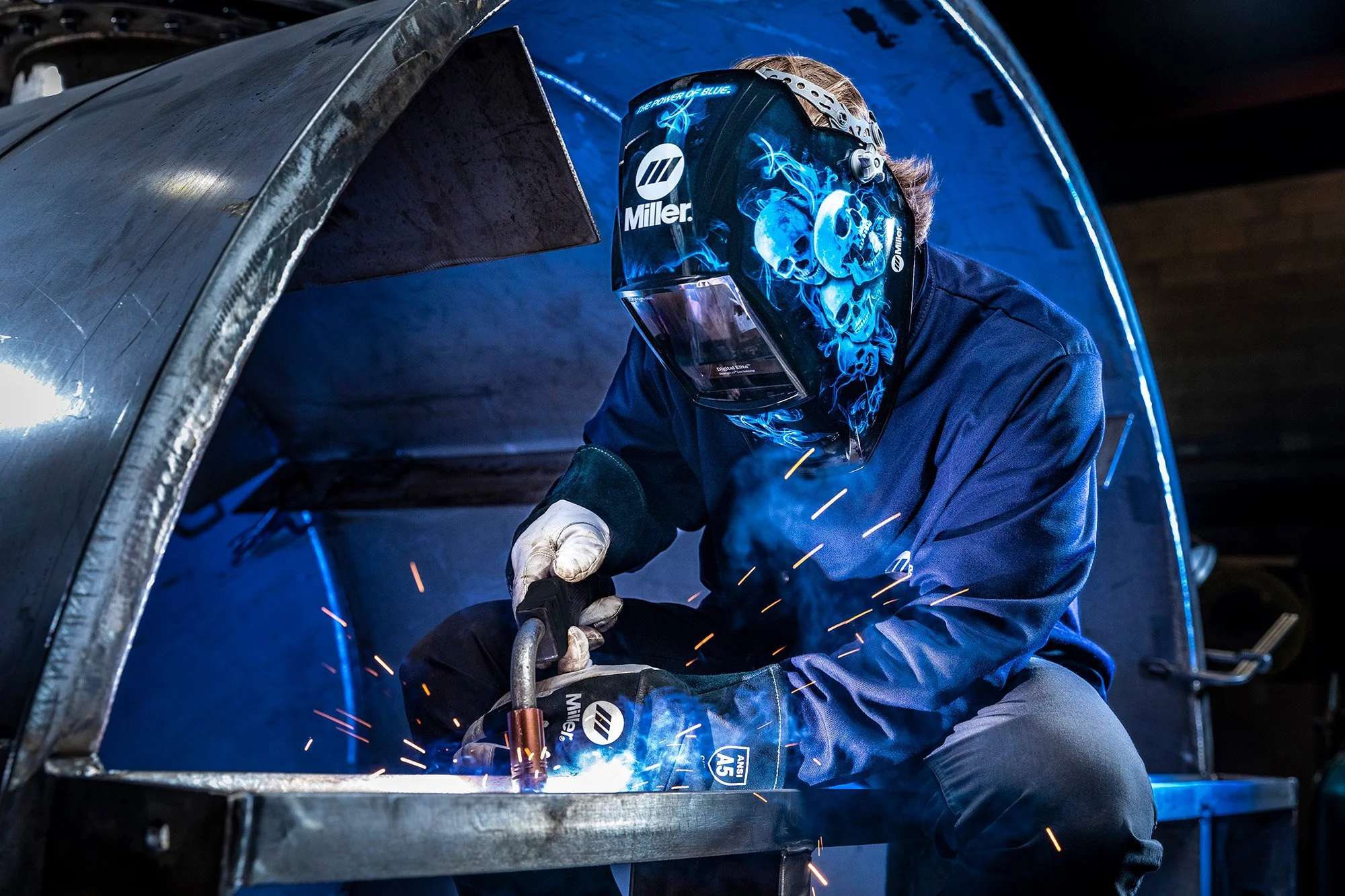 A person welding metal in a workshop, wearing a dark blue welding helmet with skulls and blue flames, and protective gloves.