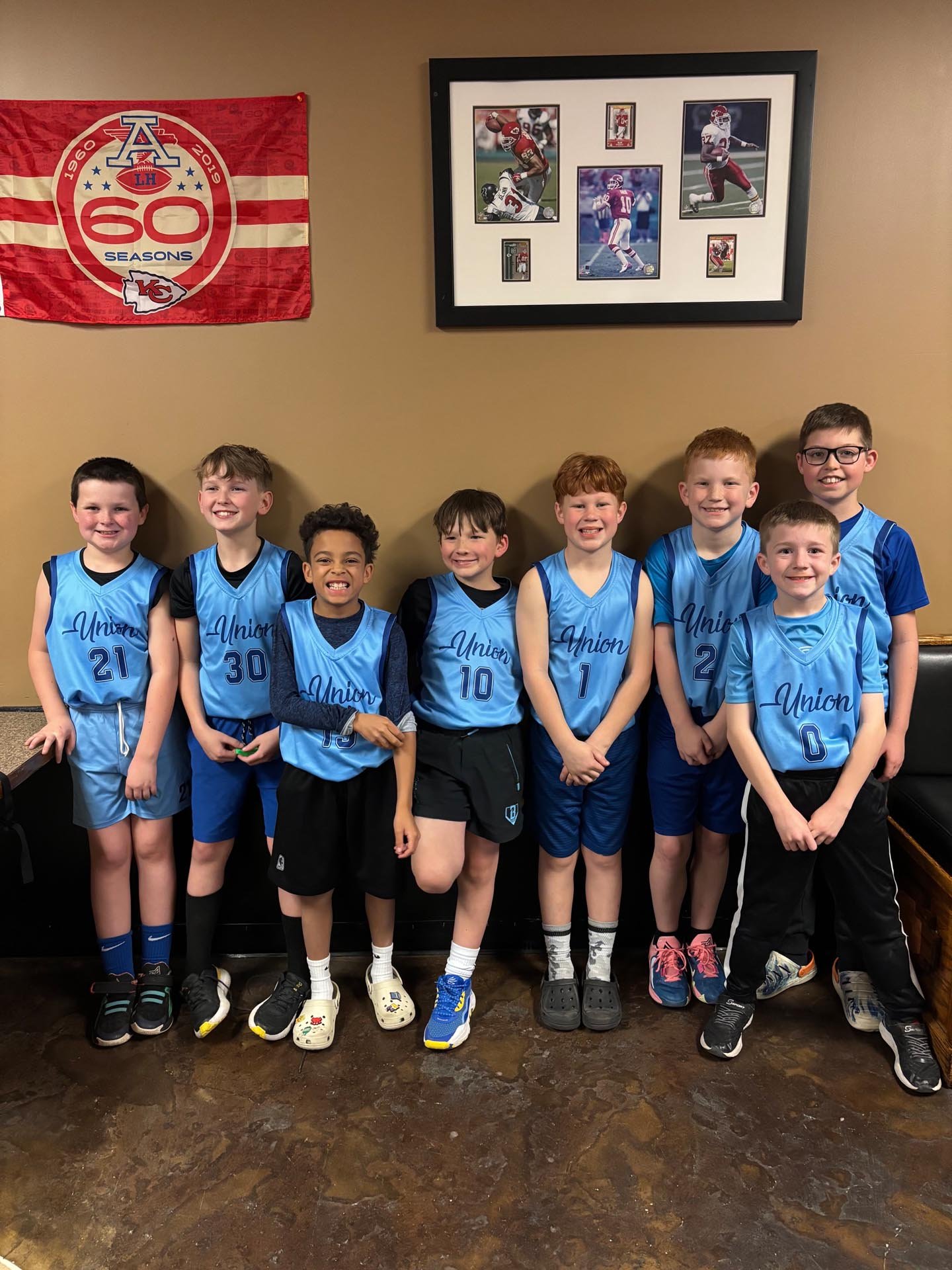 A group of seven young boys wearing blue basketball jerseys standing indoors against a beige wall, with sports memorabilia including a framed collage of football photos and a red Kansas City Chiefs flag hanging behind them.