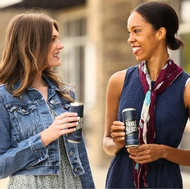 Two women smiling and talking outdoors while holding cans that say 'Noble Berry'.