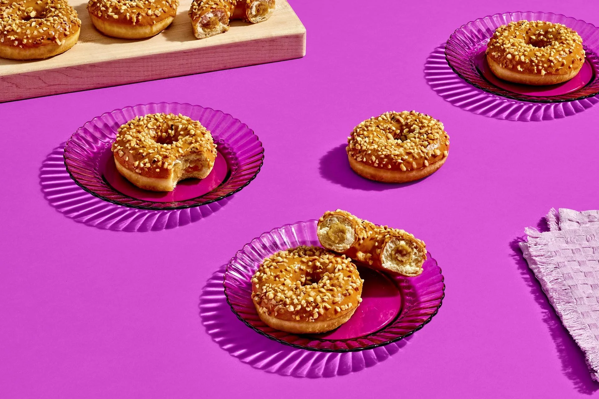 Caramel-glazed donuts with chopped nuts on pink glass plates, with a partially eaten donut revealing a caramel filling, on a pink background with a wooden board and a pink cloth.