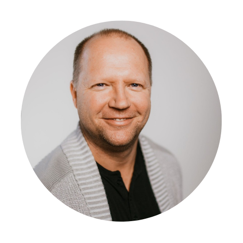 Headshot of a smiling man with a light gray cardigan, black shirt, and short hair, against a light gray background.