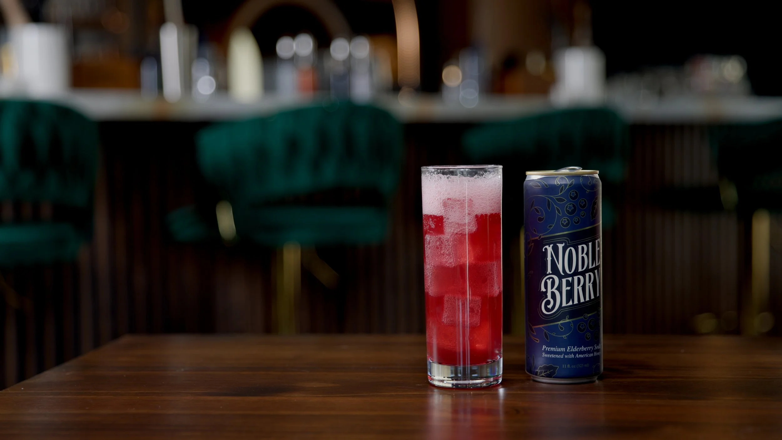 Glass of red beverage next to a can of Noble Berry Elderberry soda on a wooden table.