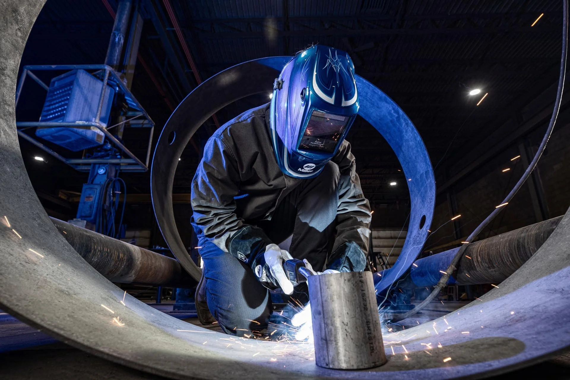A person wearing a welding helmet and gloves welds a large metal pipe in an industrial setting, with sparks flying from the welding process.