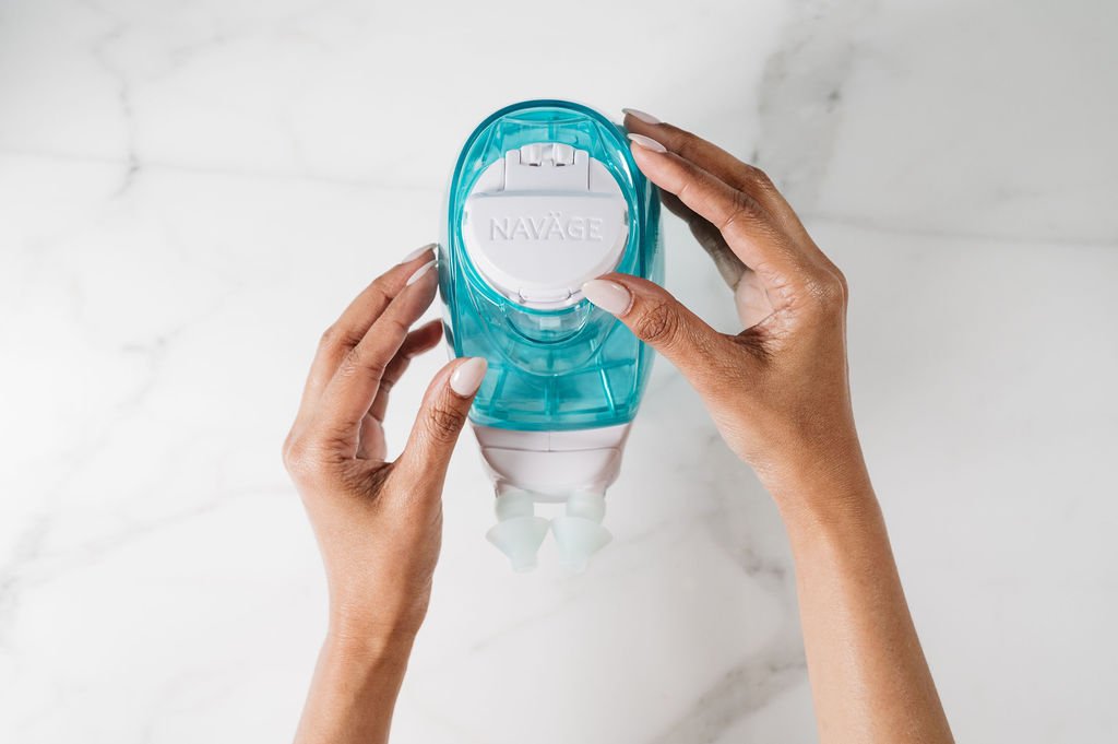 Hands holding a blue and white Naväge nasal irrigation device over a white marble countertop.