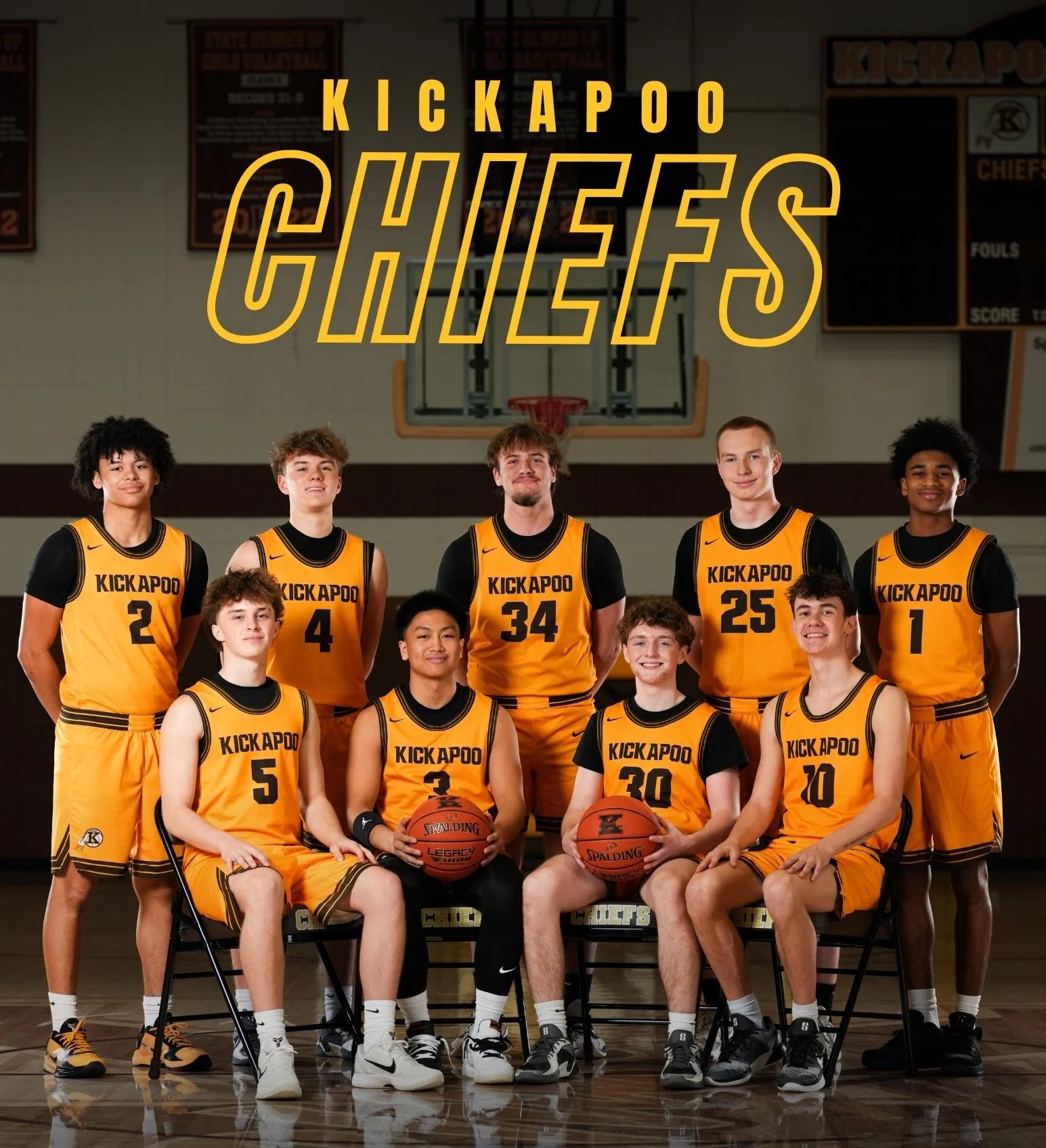 A group of young male basketball players in yellow uniforms with 'Kickapoo' written on them, posing for a team photo in a gymnasium. Some players are seated holding basketballs, and others are standing behind them. The background features a basketball hoop and scoreboard.