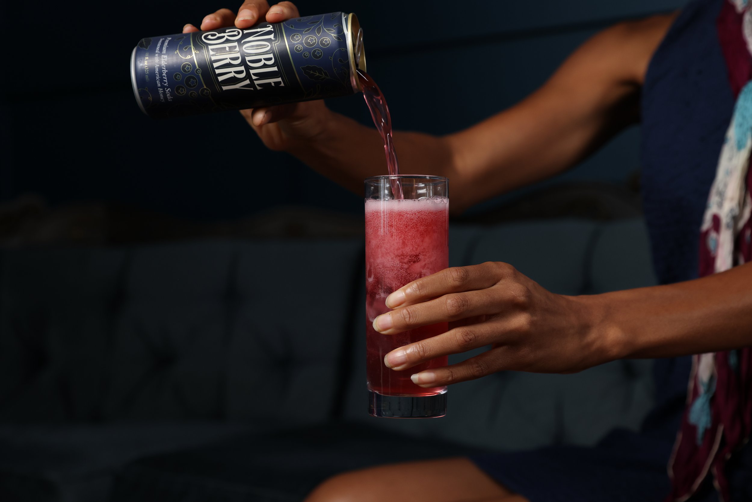 A person is pouring a pinkish-red beverage from a canned Neon Black Cherry soda into a tall glass, with only their hand and part of their arm visible.