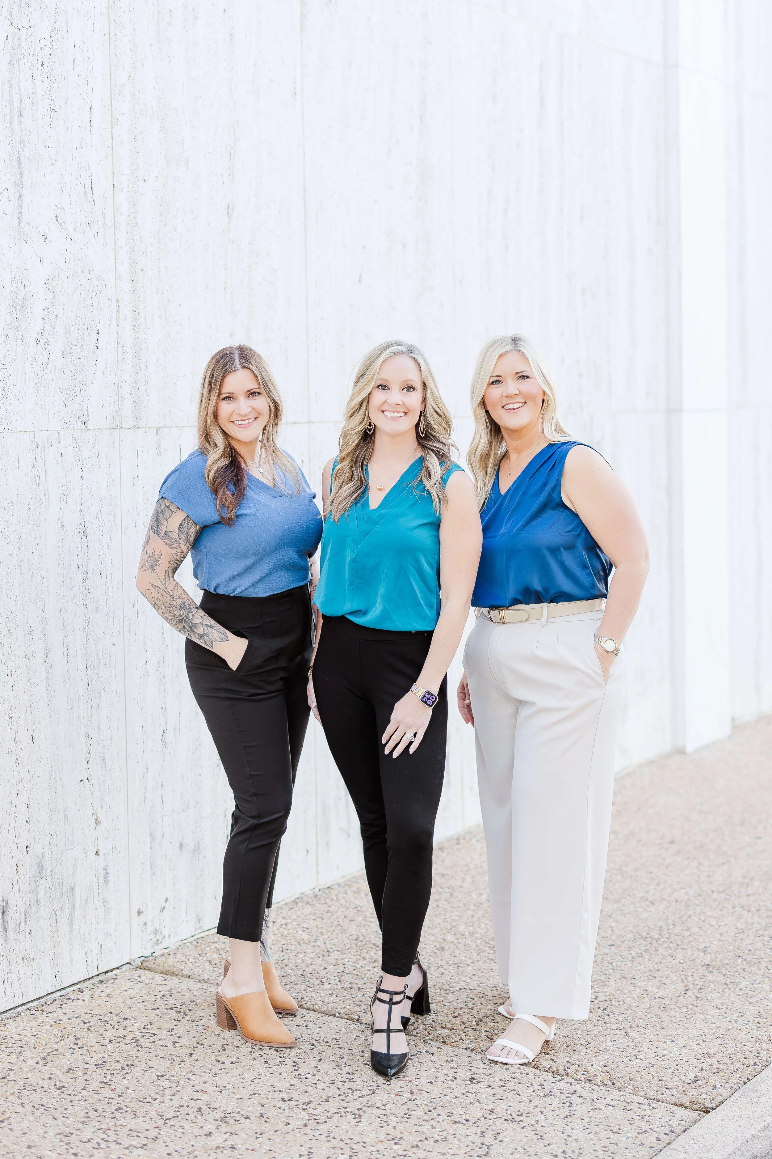 Three women standing close together outside against a white concrete wall, smiling at the camera. They are dressed in smart casual attire with various pants and tops, and wearing different styles of shoes. One woman has tattoos on her arm, and all have blonde or light brown hair.
