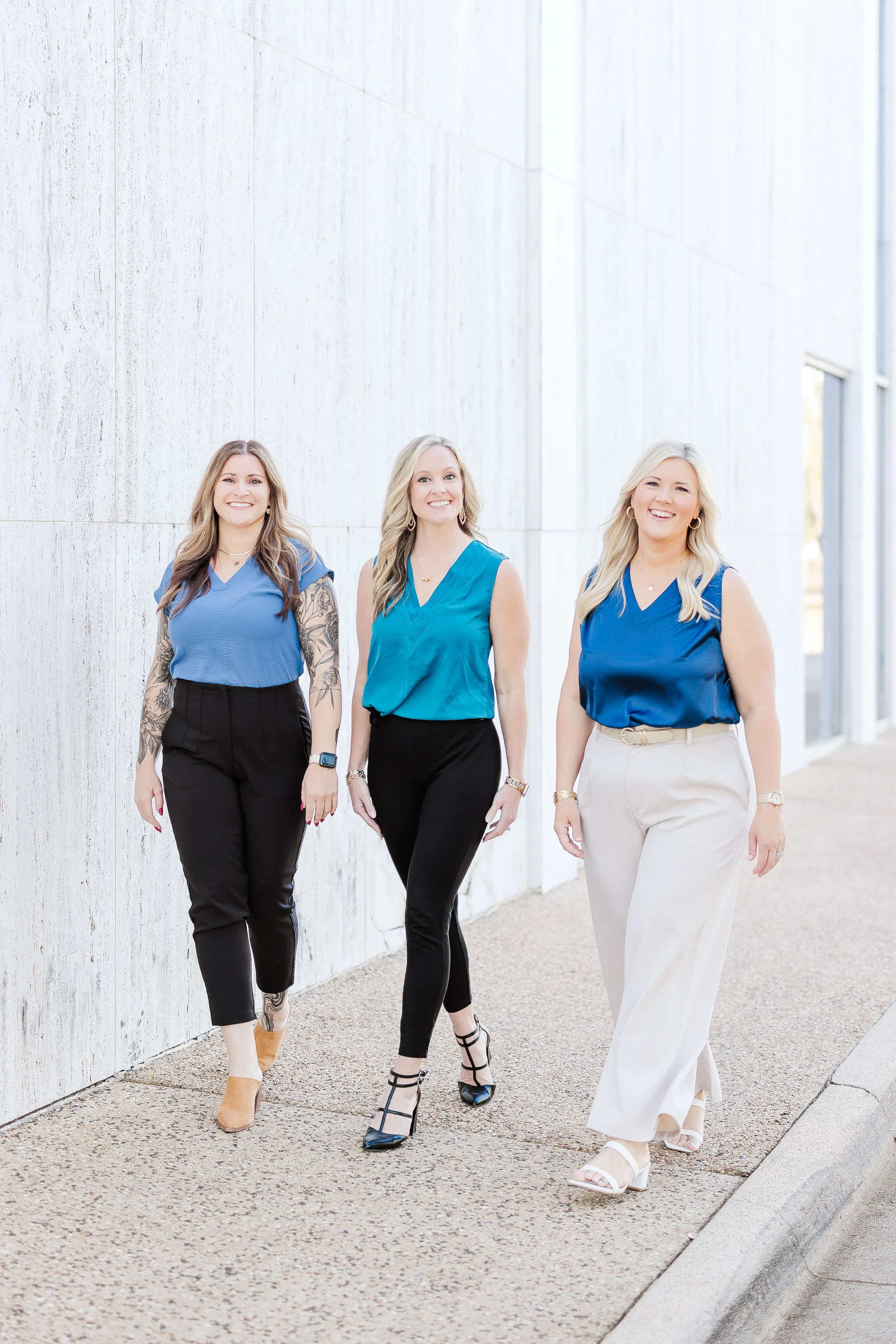 Three women walking outdoors along a sidewalk, smiling, dressed in blue tops and black or light-colored pants, near a modern white building with large windows.