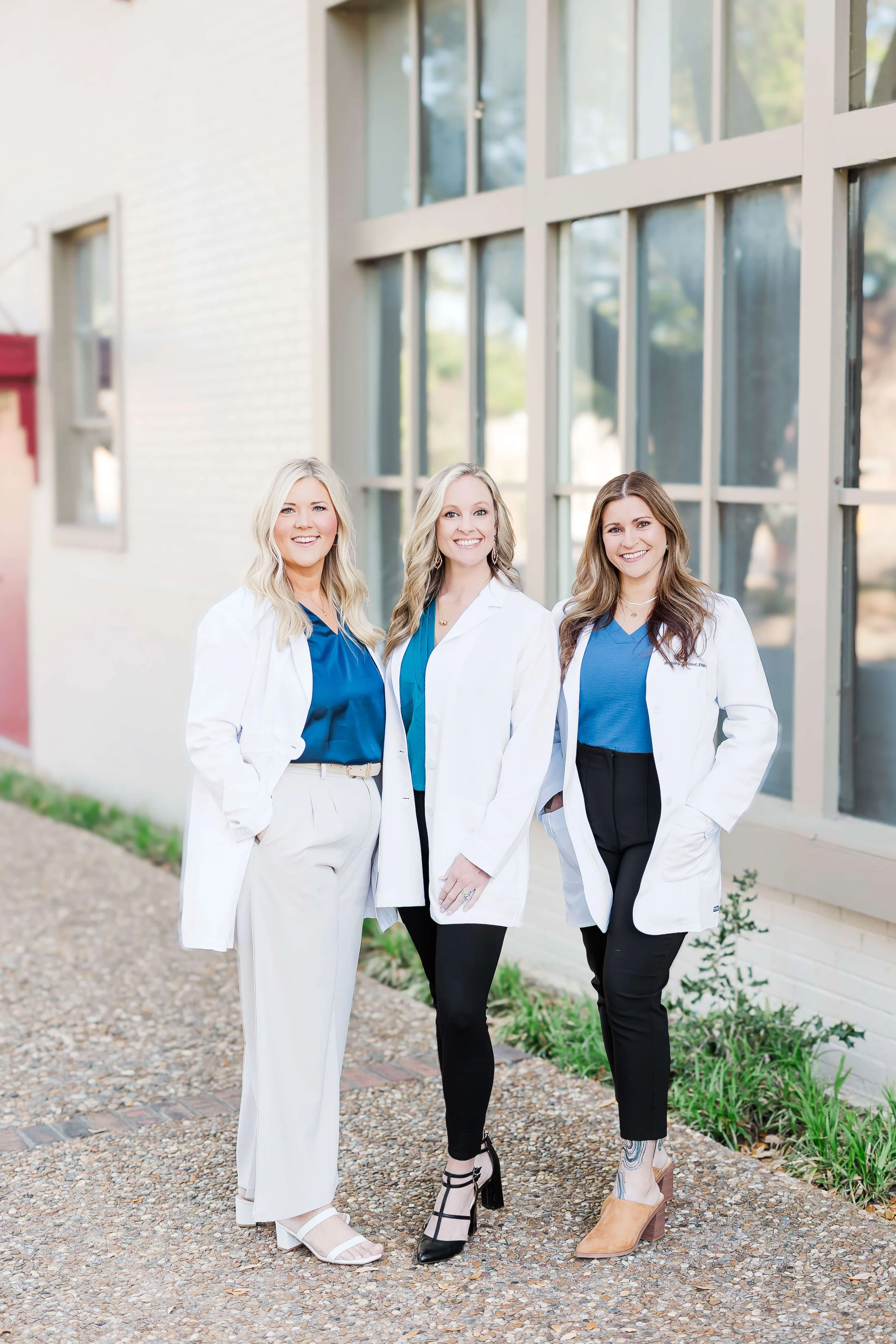 Three women dressed in professional attire standing outside in front of a building with large windows, smiling at the camera.