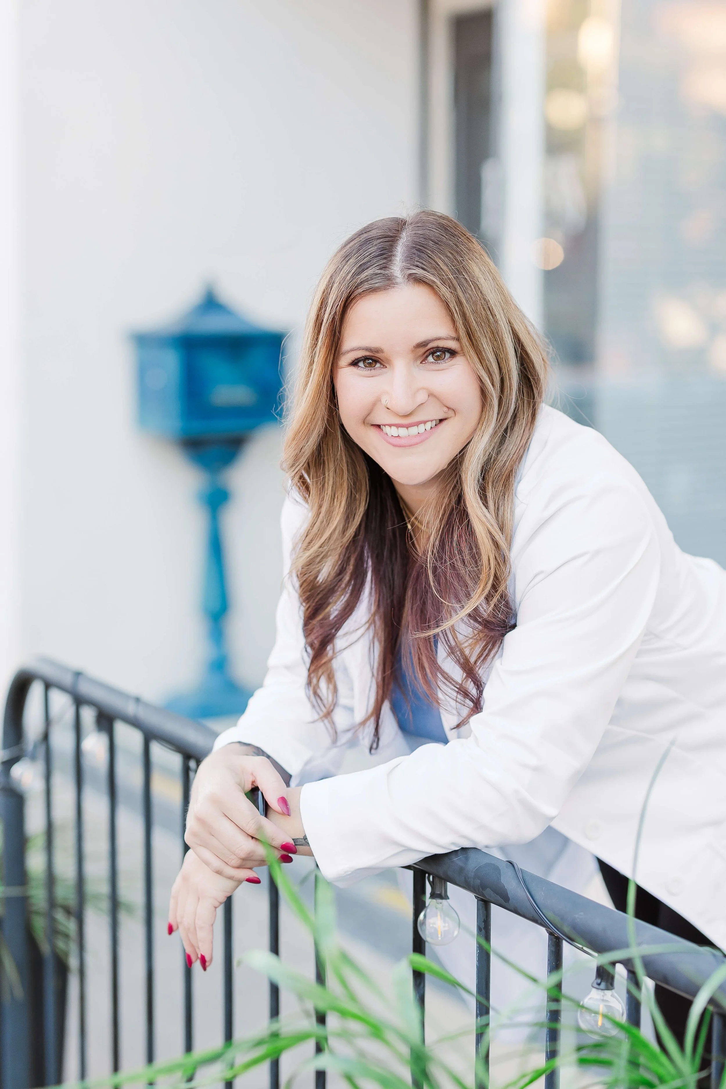 A young woman with long, wavy, light brown hair and a nose piercing, smiling and leaning on a black railing outdoors, with a large window and blurred cityscape in the background.