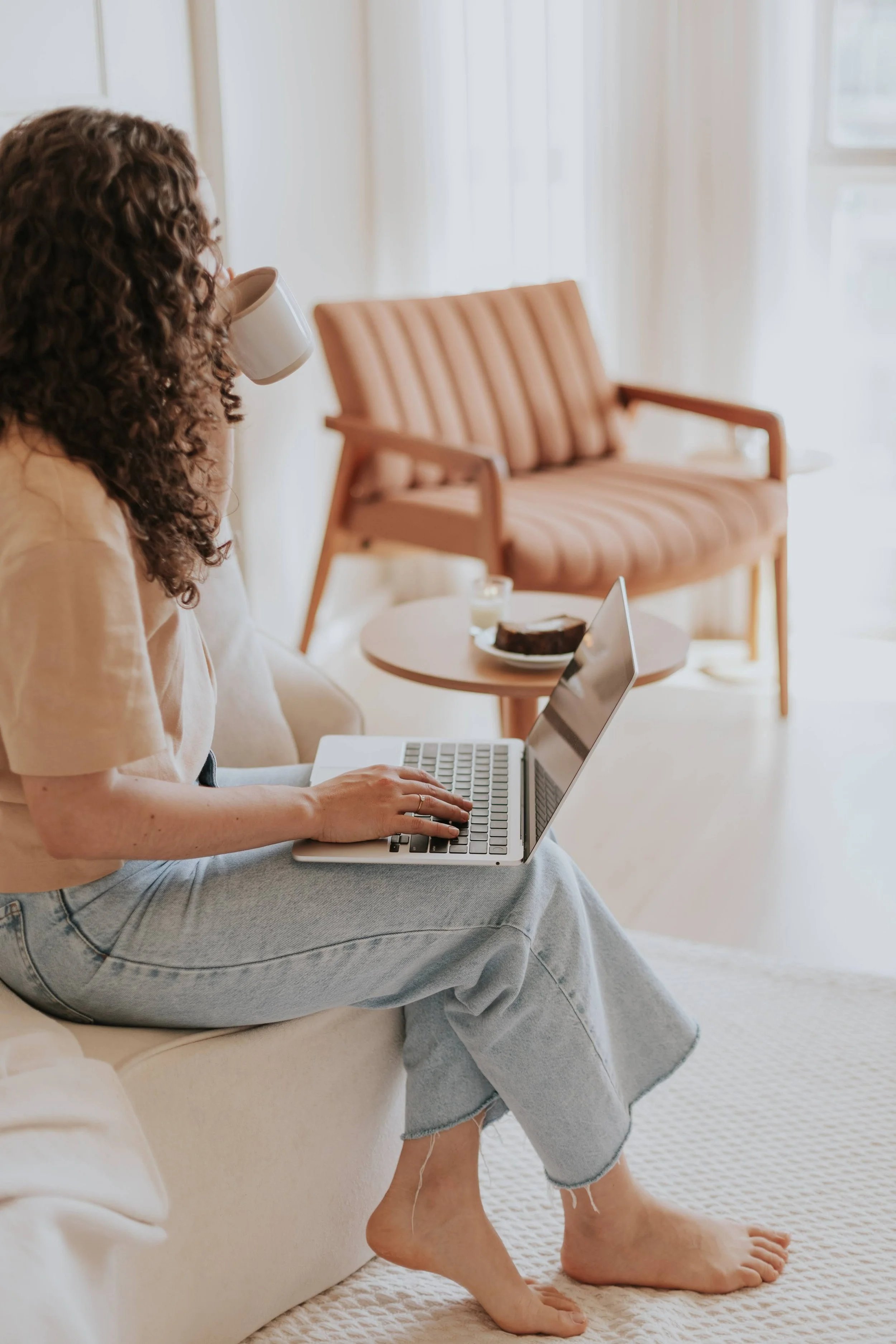 A person with curly hair sitting on a beige sofa, working on a laptop. They are wearing a beige top and gray jeans. Their left hand is on the keyboard, and their right hand is near the touchpad. They are drinking from a white mug. In the background, there is a peach-colored sofa, a small round table with a cake, a candle, and a laptop, and white curtains letting in natural light.
