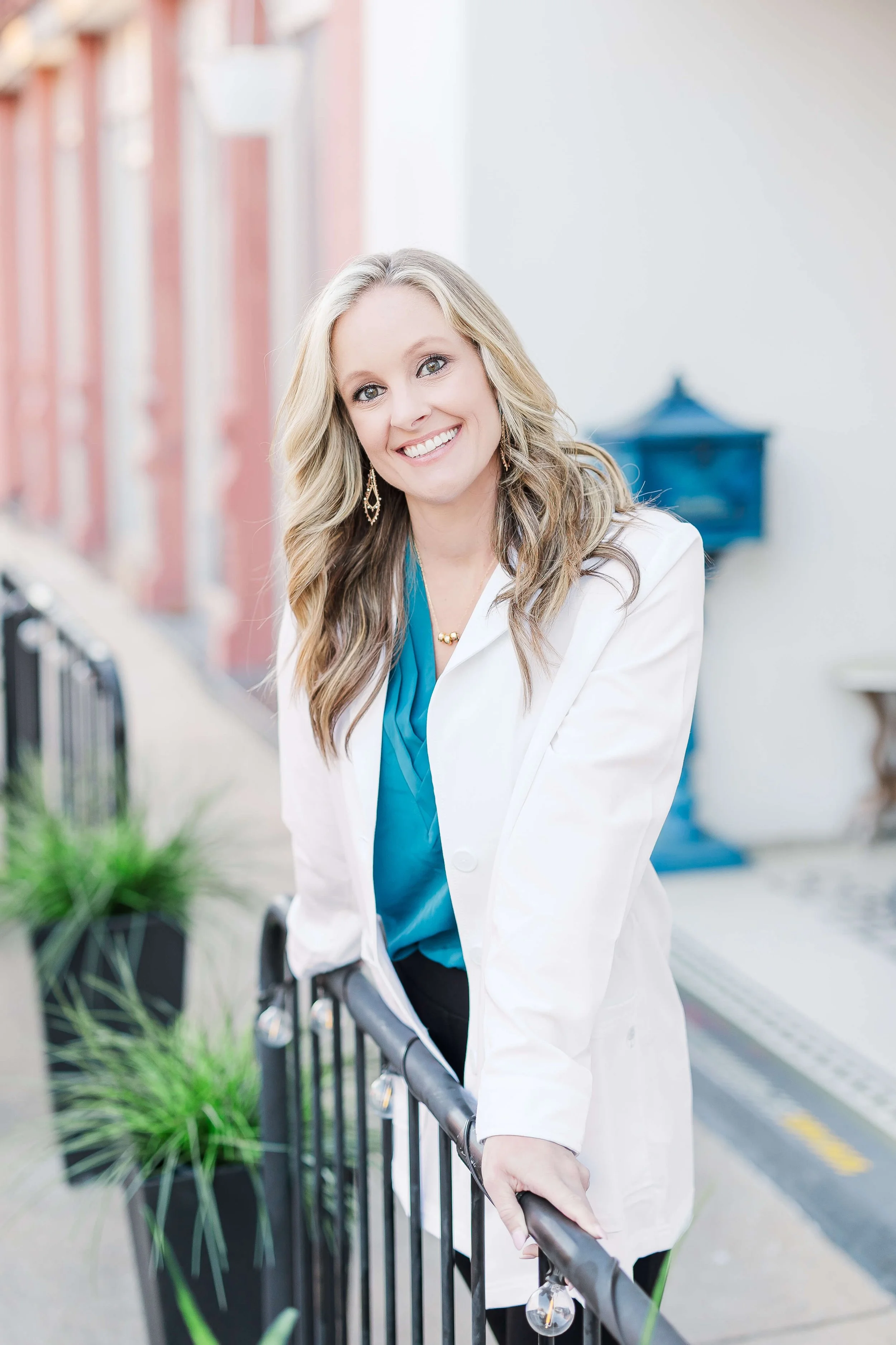Portrait of a smiling woman with long blonde hair, wearing a white blazer over a blue top, standing outdoors near a black metal fence with green plants, in an urban setting with a building and a blue mailbox in the background.