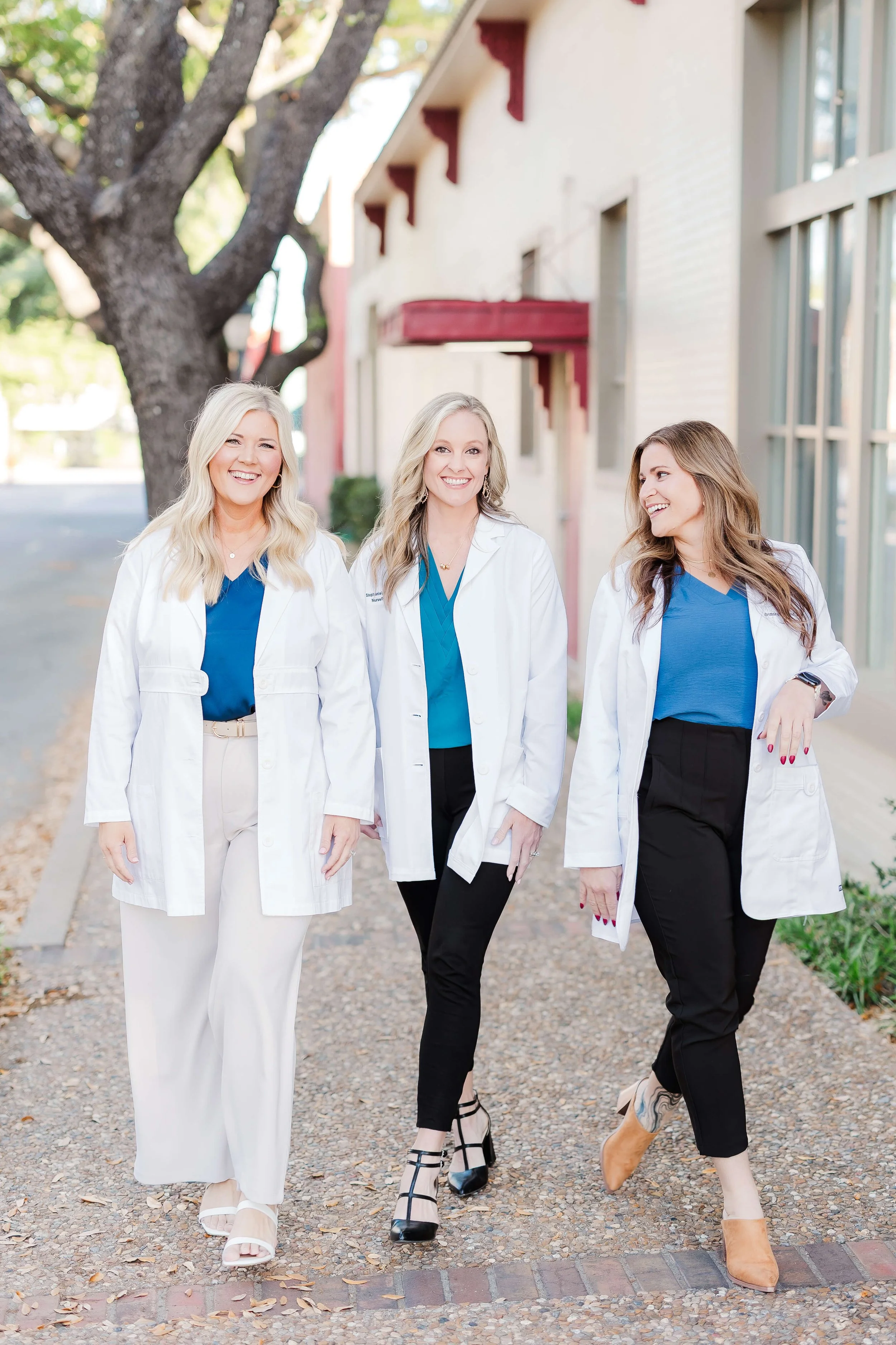 Three women in white lab coats walking outdoors, smiling and talking, with a large tree and building behind them.