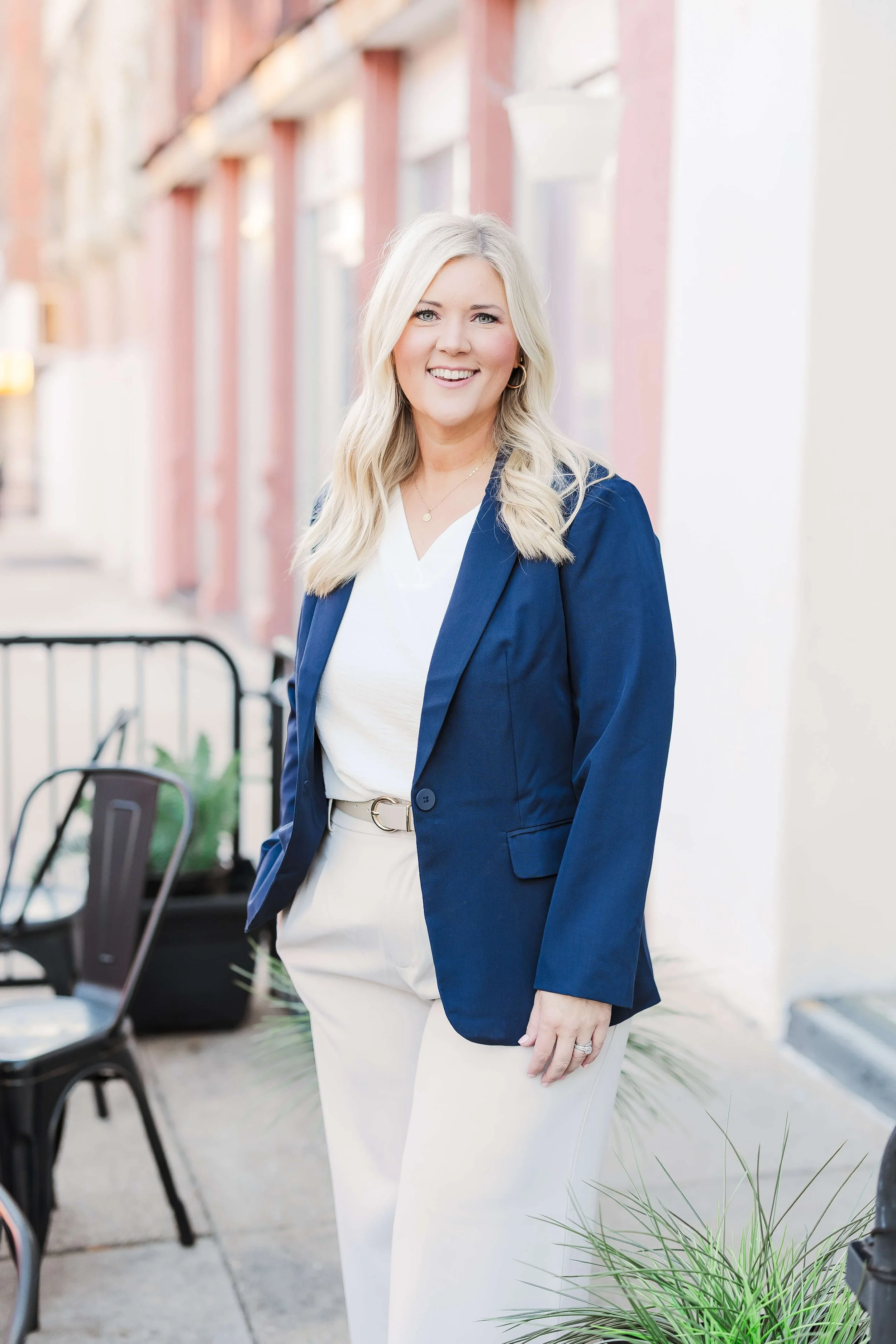 A confident blonde woman in a white dress and blue blazer standing outdoors in front of a row of buildings.
