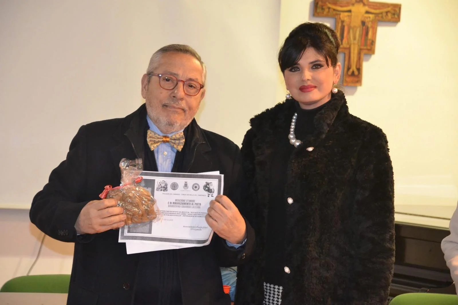 Un uomo con occhiali, giacca nera e papillon a quadri tiene una certificato e un pacchetto di biscotti, accanto a una donna con abbigliamento nero e collana di perle, in una stanza con decorazioni.