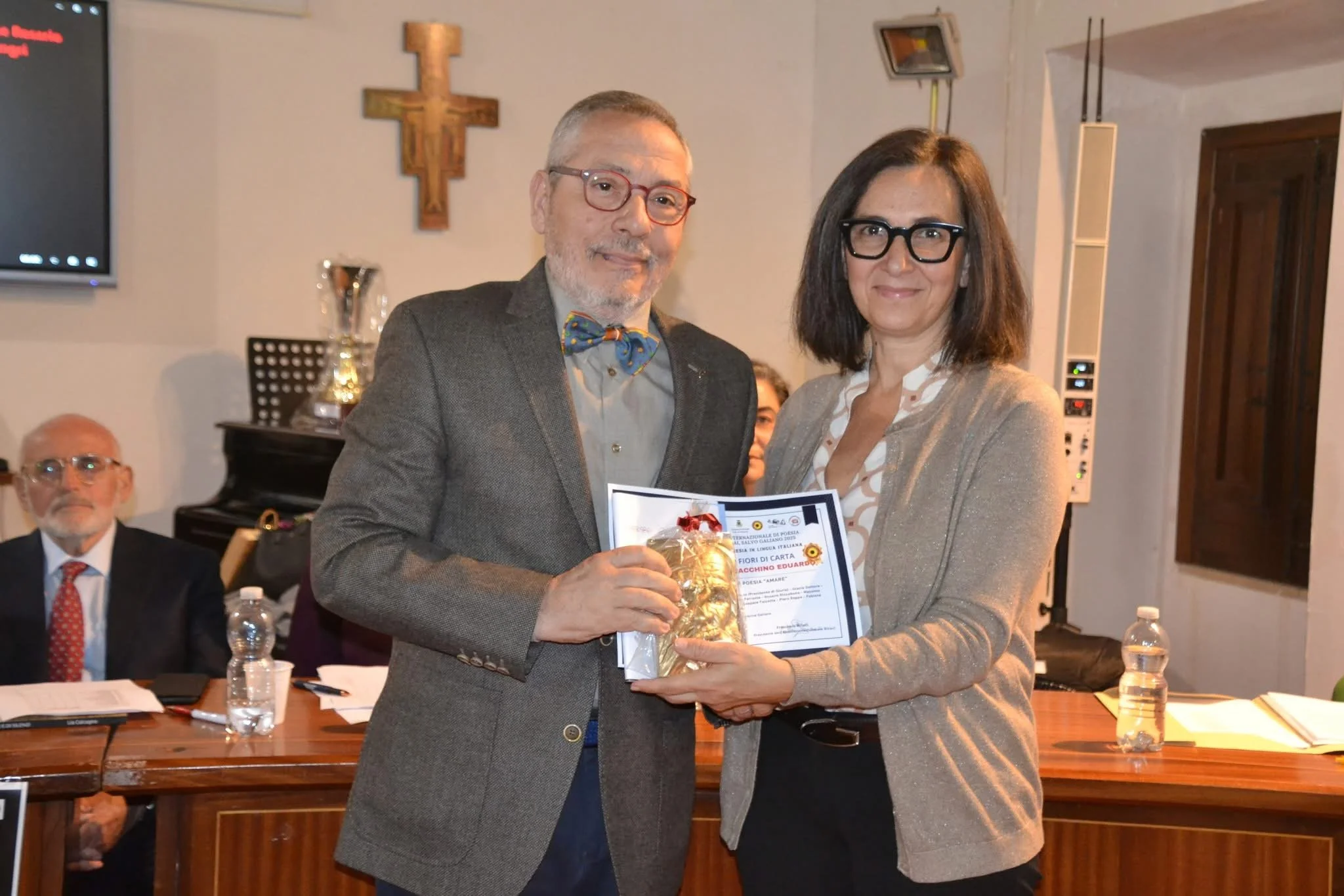 Due persone sorridenti, un uomo con occhiali rossi e una donna con occhiali neri, si scambiano un premio o un riconoscimento durante un evento formale, con altre persone sedute dietro di loro.