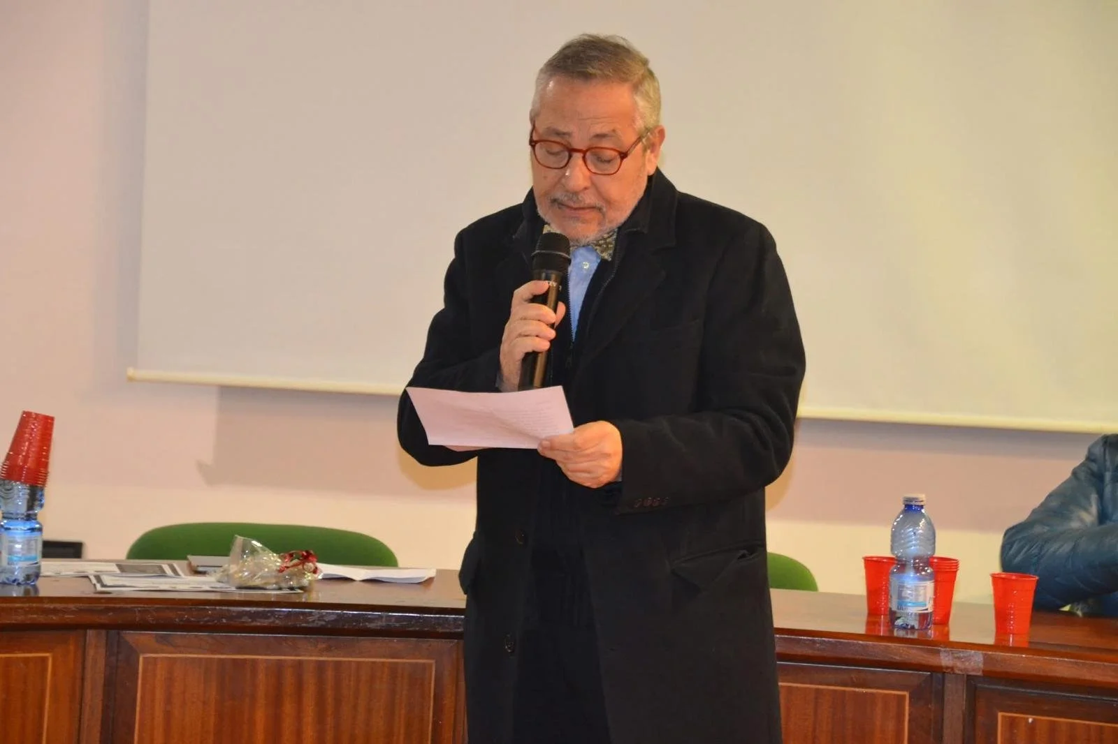 Uomo anziano con occhiali che legge un discorso o una presentazione mentre tiene in mano un microfono, in un ambiente interno con tavoli, bottiglie d'acqua, bicchieri rossi e documenti.