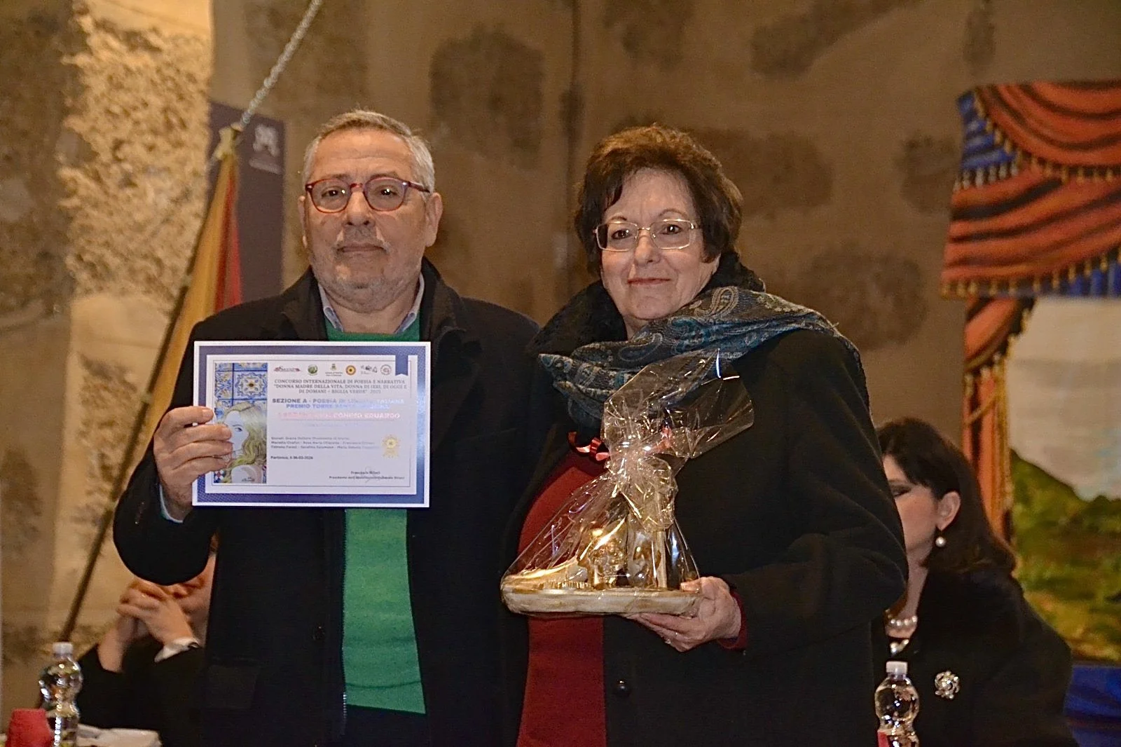 Due persone in piedi, uomo che mostra un certificato e donna con regali, in un ambiente interno durante un evento.