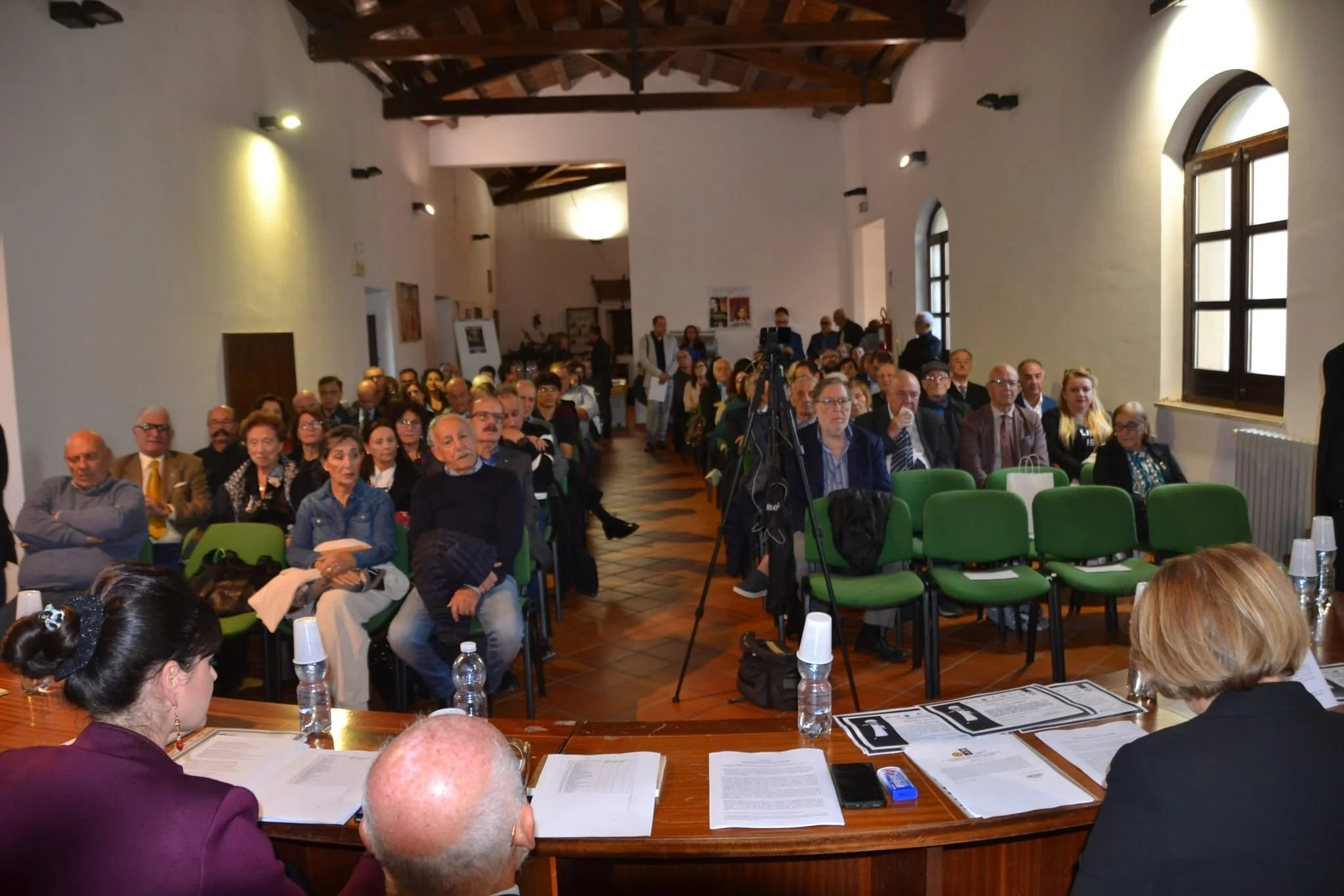 Un gruppo di persone sedute in una sala durante un evento o una conferenza, con alcuni alla testa di un tavolo e altri seduti su sedie verdi in fila, con grandi finestre e un soffitto in legno.
