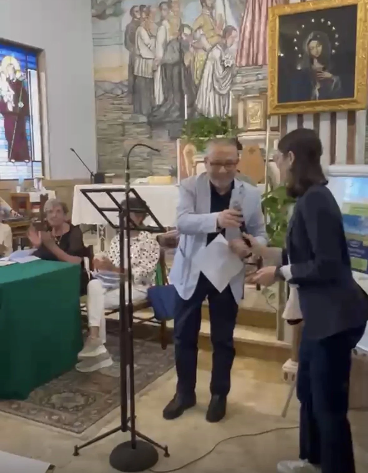 Persone che partecipano a un evento in una chiesa, con dipinti religiosi alle pareti. Due persone sono al centro, una che riceve un riconoscimento o un premio mentre l'altra gli porge qualcosa, con altre persone sedute sullo sfondo.