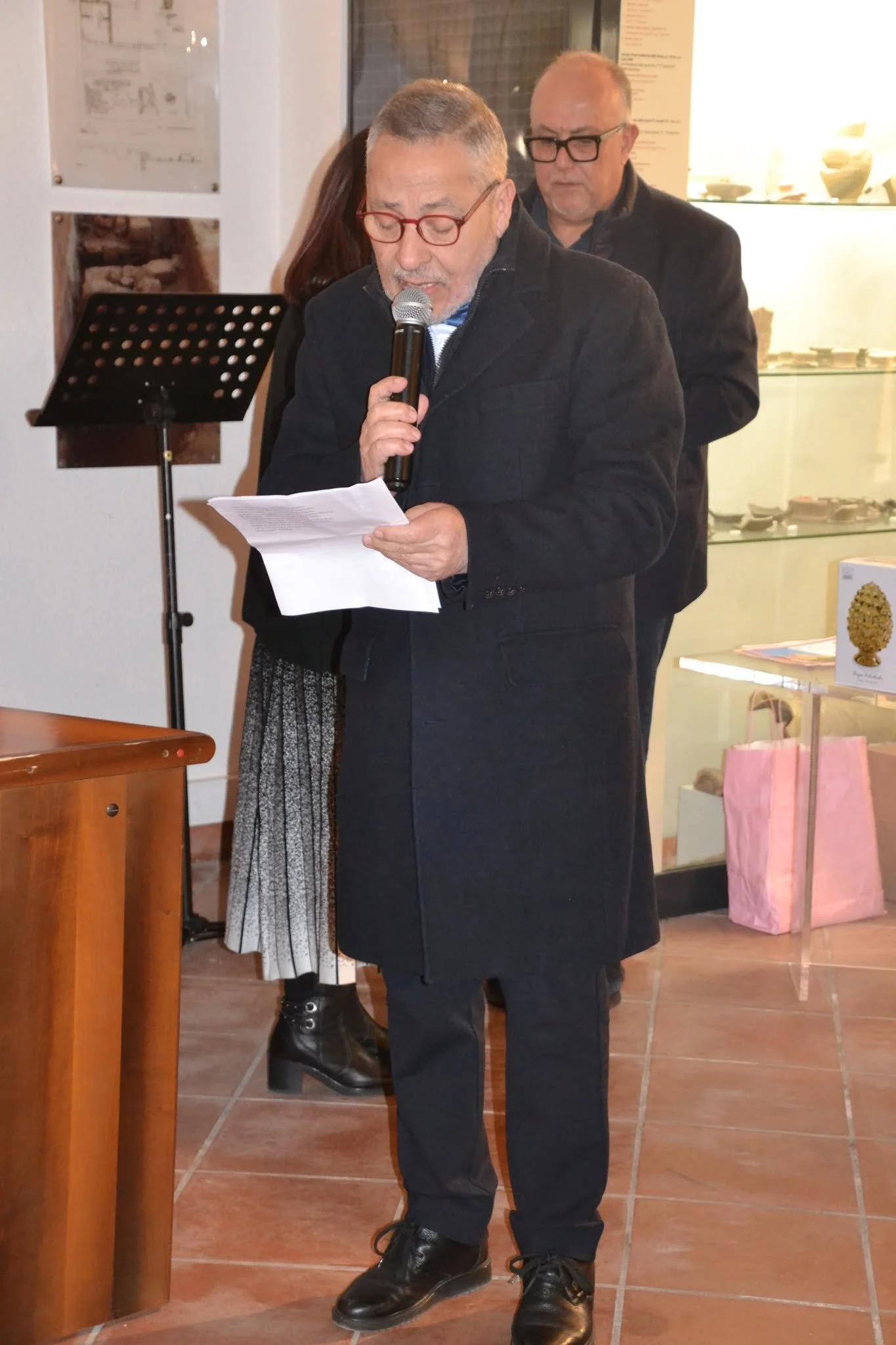 Uomo anziano con barba e occhiali rossi che parla al microfono, con altri due persone sullo sfondo in un ambiente interno, pare essere un evento o una presentazione.
