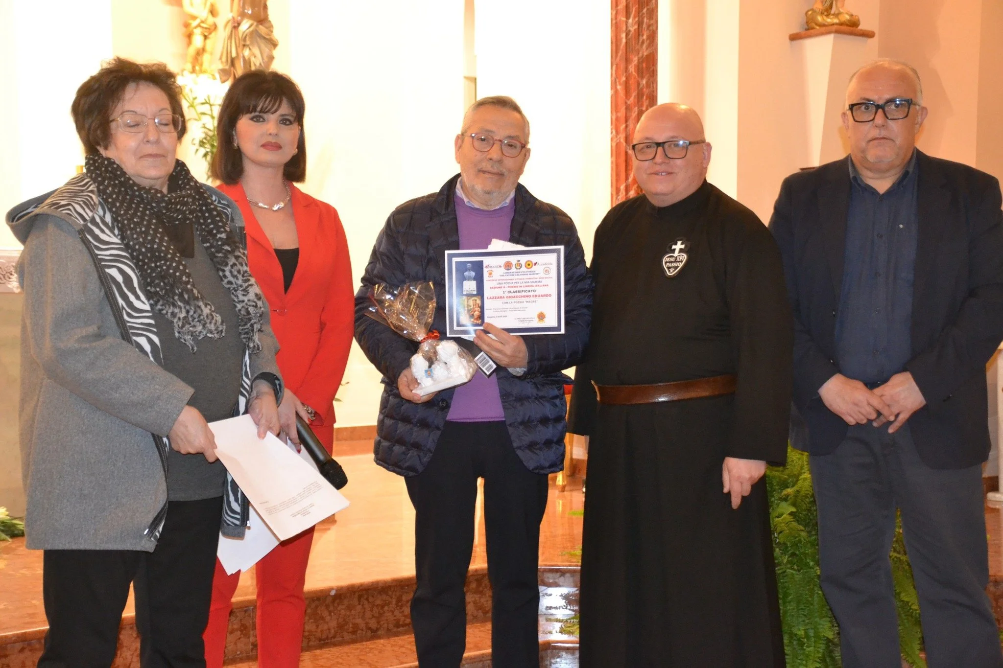 Gruppo di cinque persone durante una cerimonia, una delle quali riceve un premio o riconoscimento, con certificato e fiore, in un luogo interno decorato con statue e tende rosse.