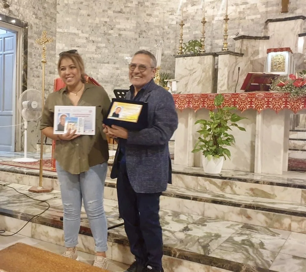 Due persone sorridenti in piedi in una chiesa o una cappella, con uno dei due che tiene un riconoscimento o una certificazione, e l'altro che tiene un poster con una foto e del testo. Sullo sfondo ci sono altari decorati con candelabri e una pianta.
