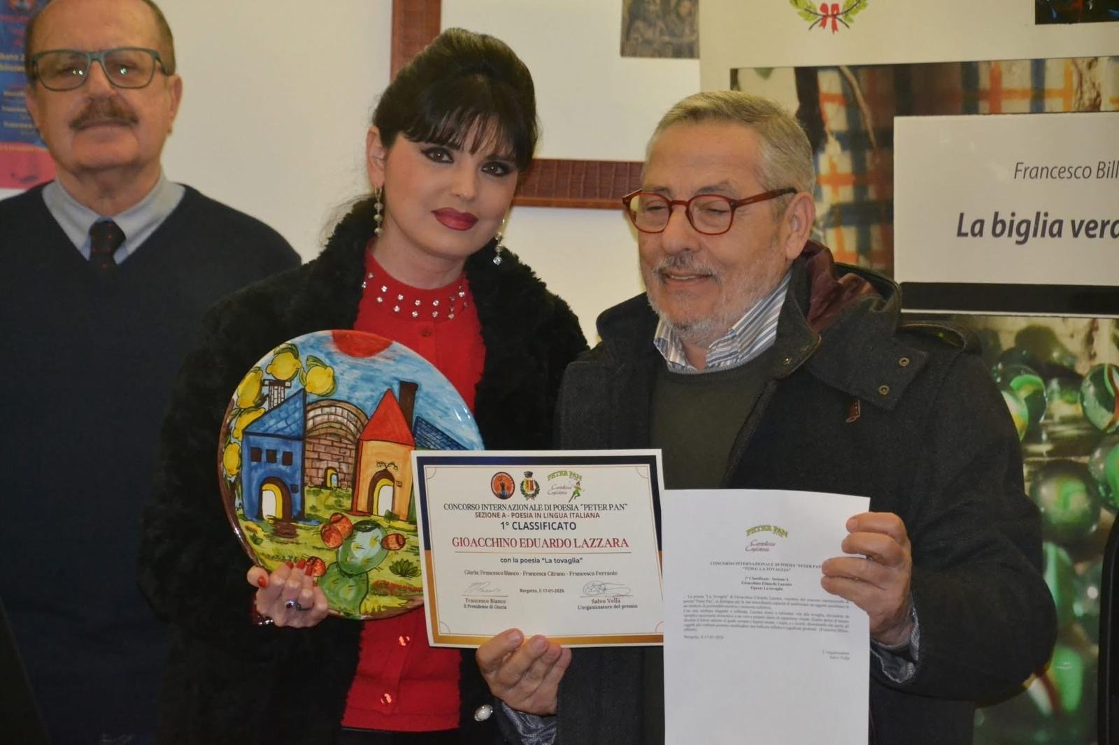 Tre persone che partecipano a un premio: una donna tiene un piatto decorato e un certificato, un uomo con occhiali e barba mostra un foglio, un uomo anziano in sfondo.