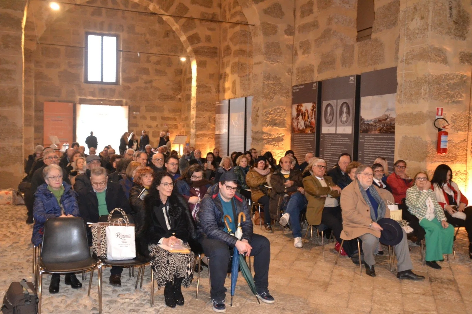 Pubblico di persone sedute che assiste a un evento in una sala con pareti di pietra e pannelli informativi, con alcuni in piedi vicino all'ingresso in background.