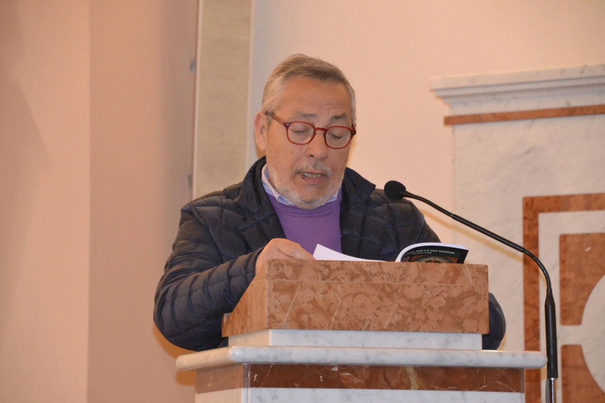 Uomo anziano con barba e occhiali rossi che legge un libro davanti a un pulpito di marmo rosa durante una presentazione o discorso in un ambiente interno.
