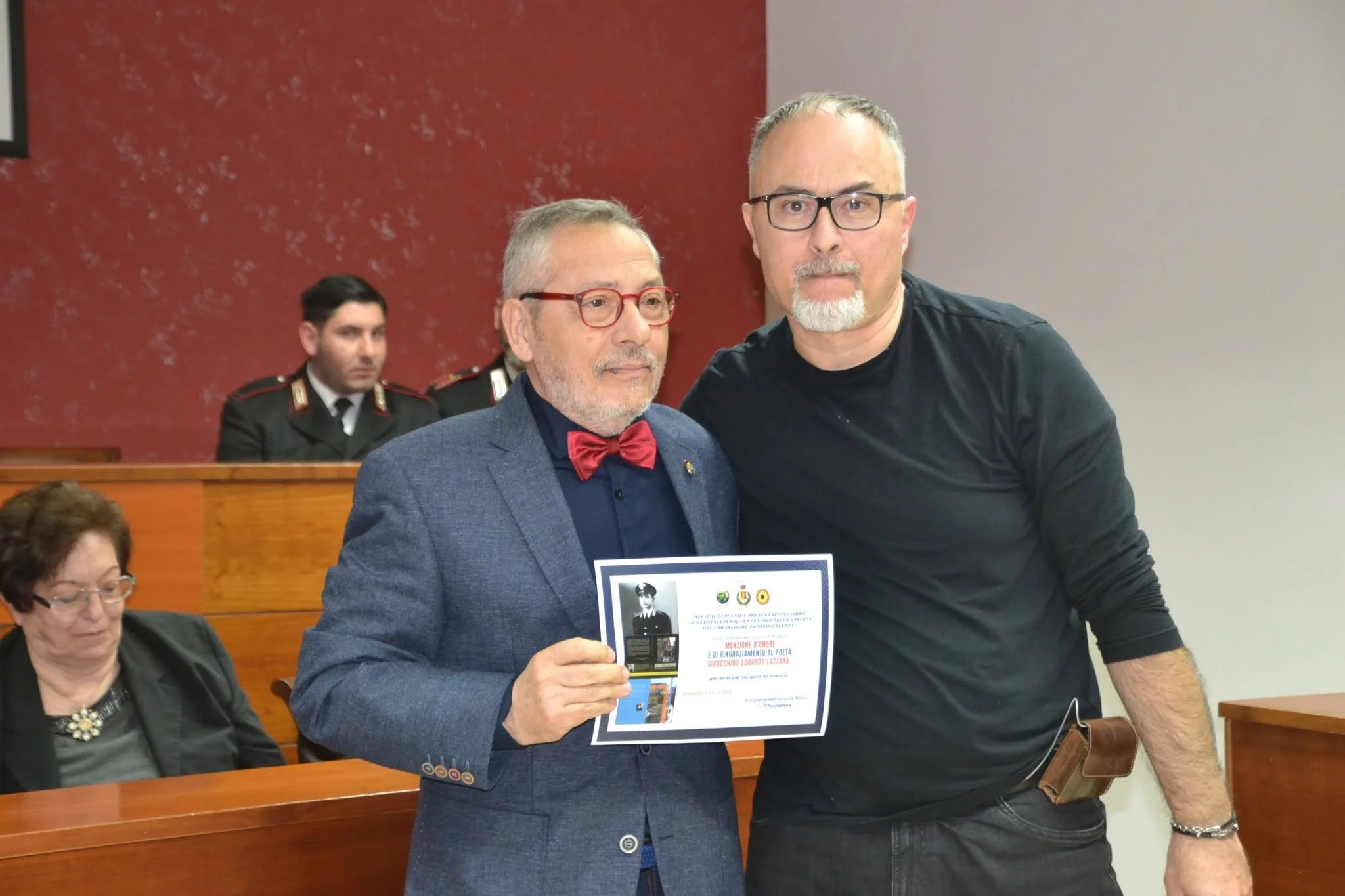 Due uomini in un evento formale, uno con barba e occhiali rettangolari che tiene un certificato e l'altro accanto con capelli corti e barba grigia, vestiti elegantemente. Sullo sfondo, due militari in uniforme sono seduti.