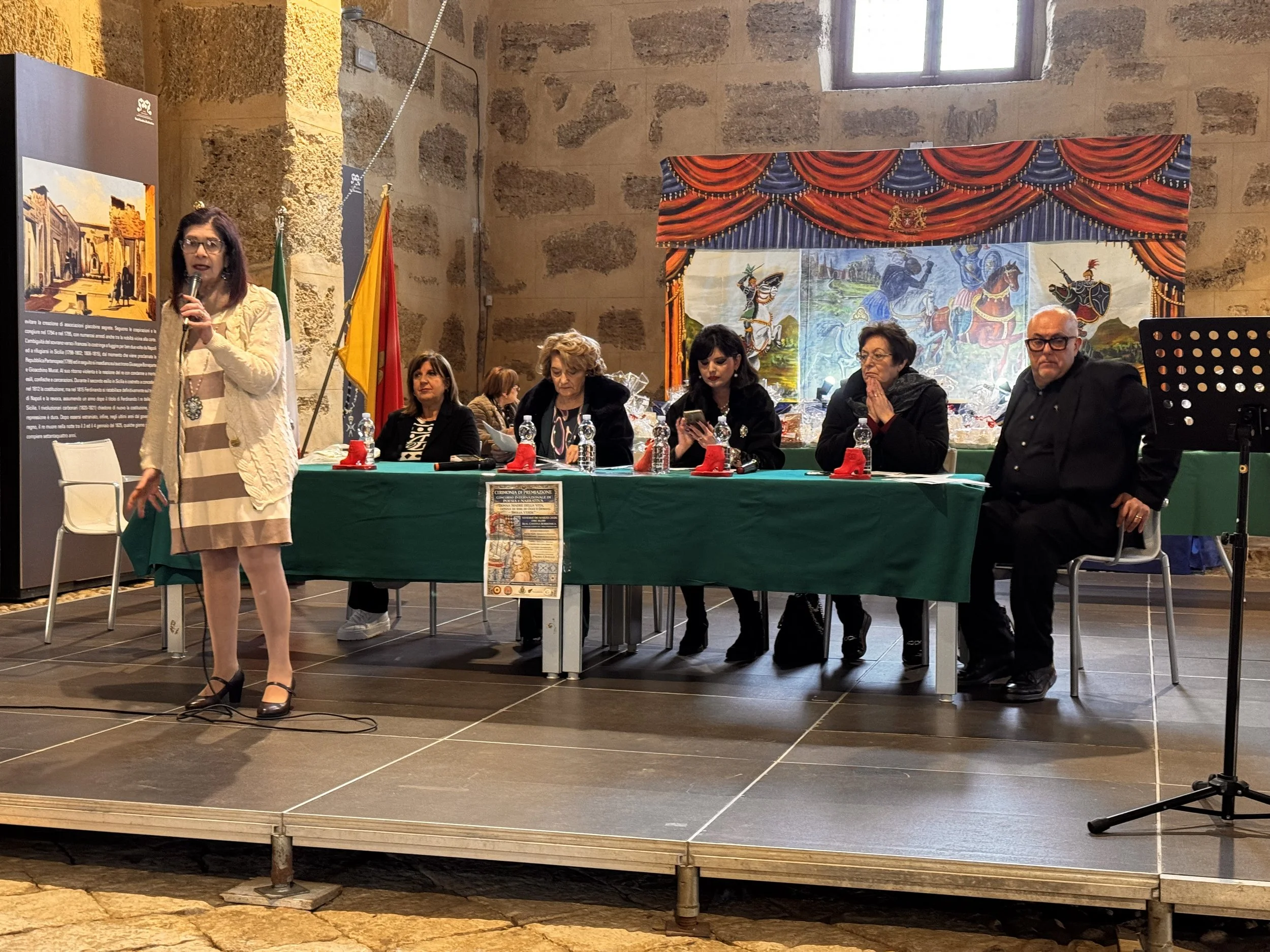Un evento culturale o conferenza con sei persone sedute dietro a un tavolo, una donna si sta avvicinando con un microfono, sullo sfondo un sipario teatrale dipinto con scene storiche e italiani, e alcune bandiere.