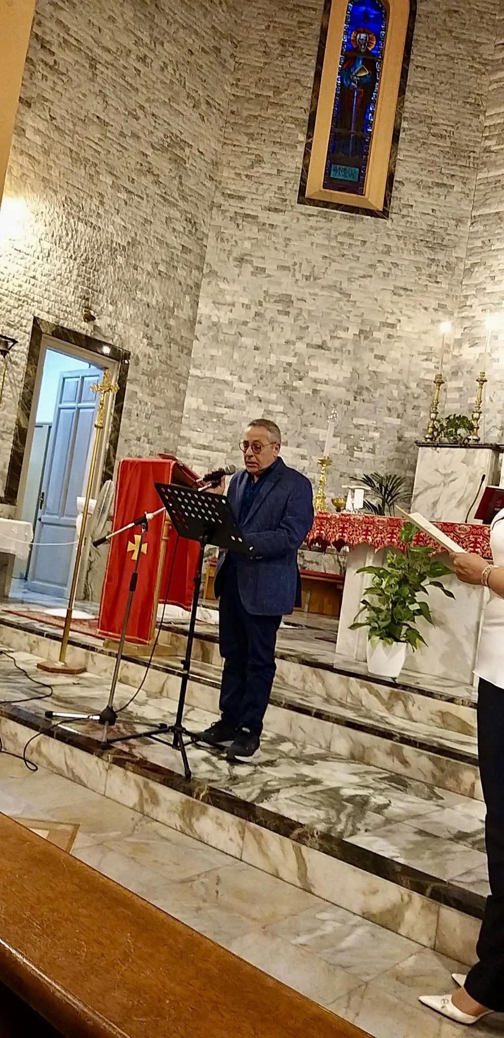 Un uomo vestito elegantemente parla in un microfono all'interno di una chiesa. Sullo sfondo, un altare decorato con tessuti rossi e candele.
