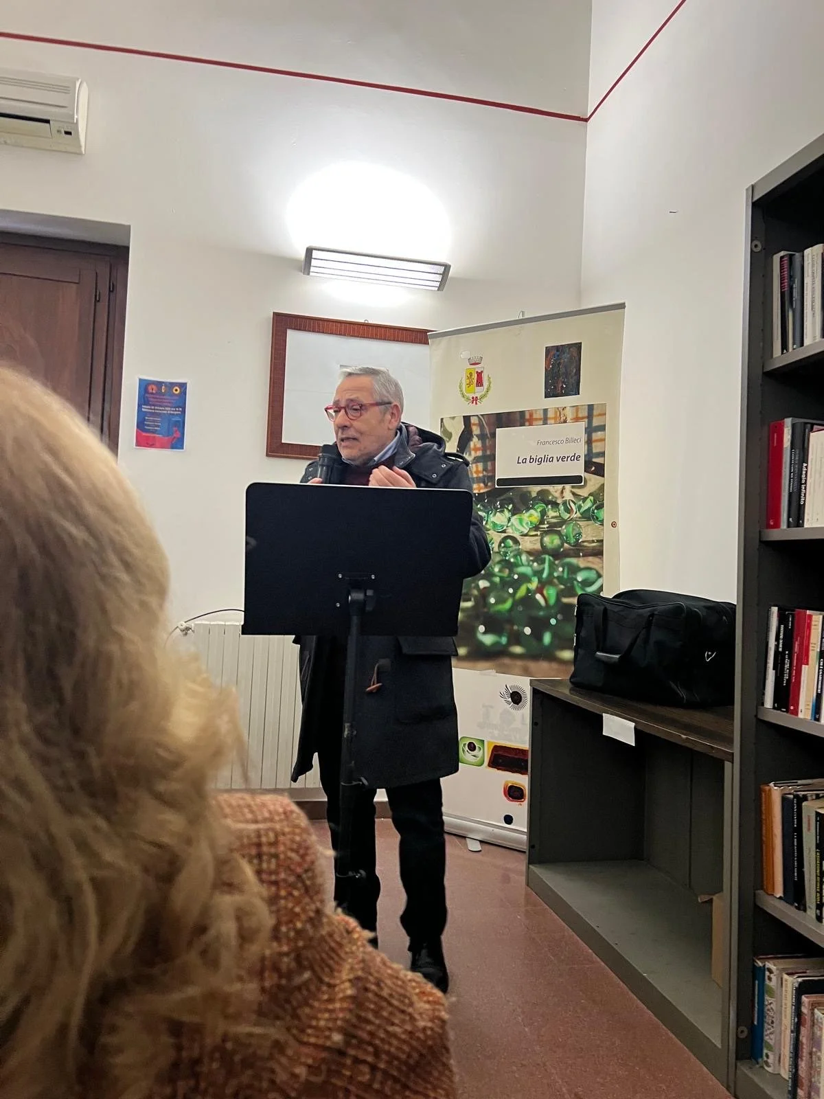 Un uomo anziano con occhiali rossi tiene un microfono e parla durante una presentazione in una libreria o sala pubblica, con un pubblico che ascolta, libreria visibile a destra e uno sfondo con una copertina di libro sulla parete.