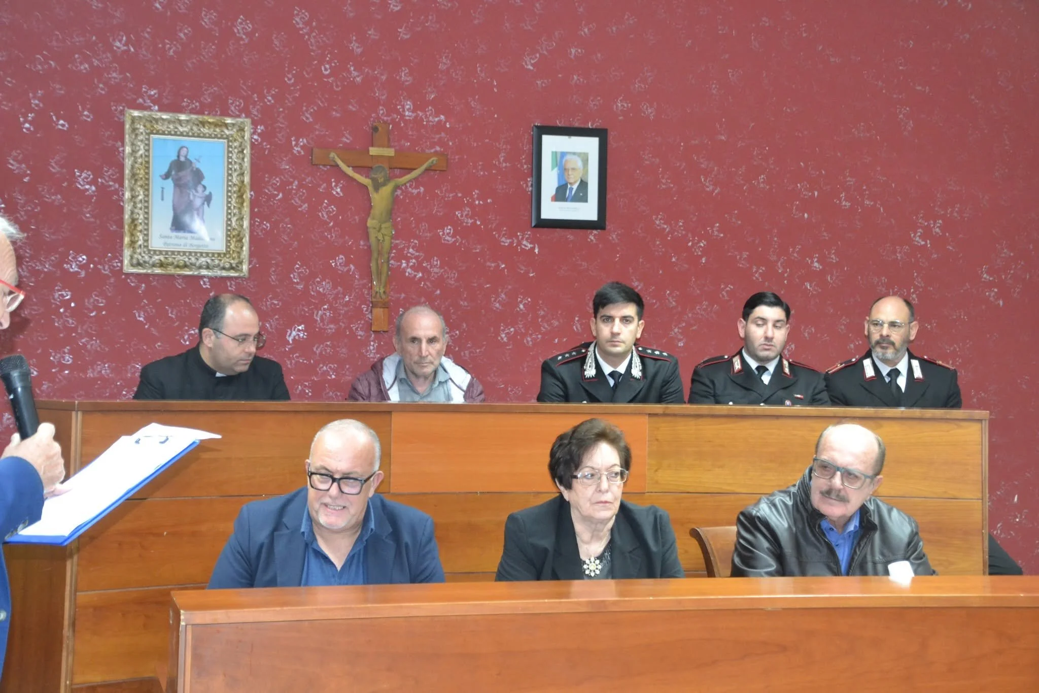 Riunione o assemblea con persone sedute dietro un banco in una stanza con pareti rosse decorate con immagini religiose, tra cui una crocifissione e un quadro di una figura umana. Alcune persone sono impegnate con documenti o in conversazioni.