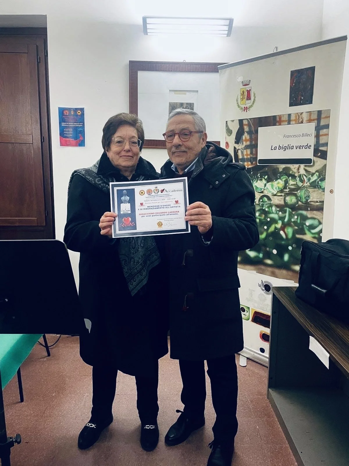 Due persone, un uomo e una donna, mostrano un certificato di riconoscimento o premio mentre posano per una foto in un luogo interno. Sullo sfondo si vede una tenda con un'immagine di un albero di vetro.