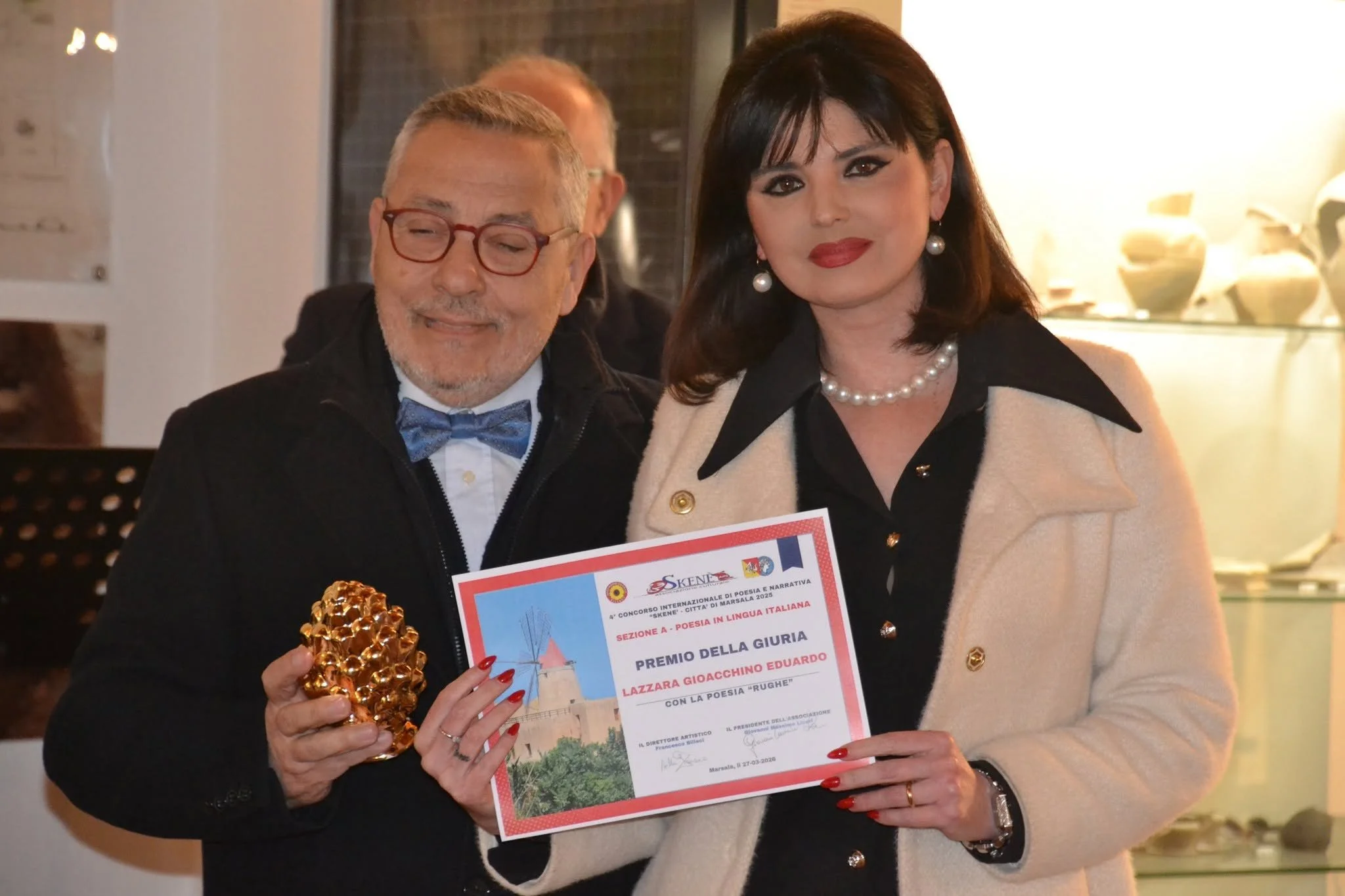 Una donna e un uomo sorridenti sono in un ambiente interno, la donna tiene un certificato e una statuetta d'oro. Sono in occasione di un premio o riconoscimento, con decorazioni eleganti aperto, e portano abiti formali.