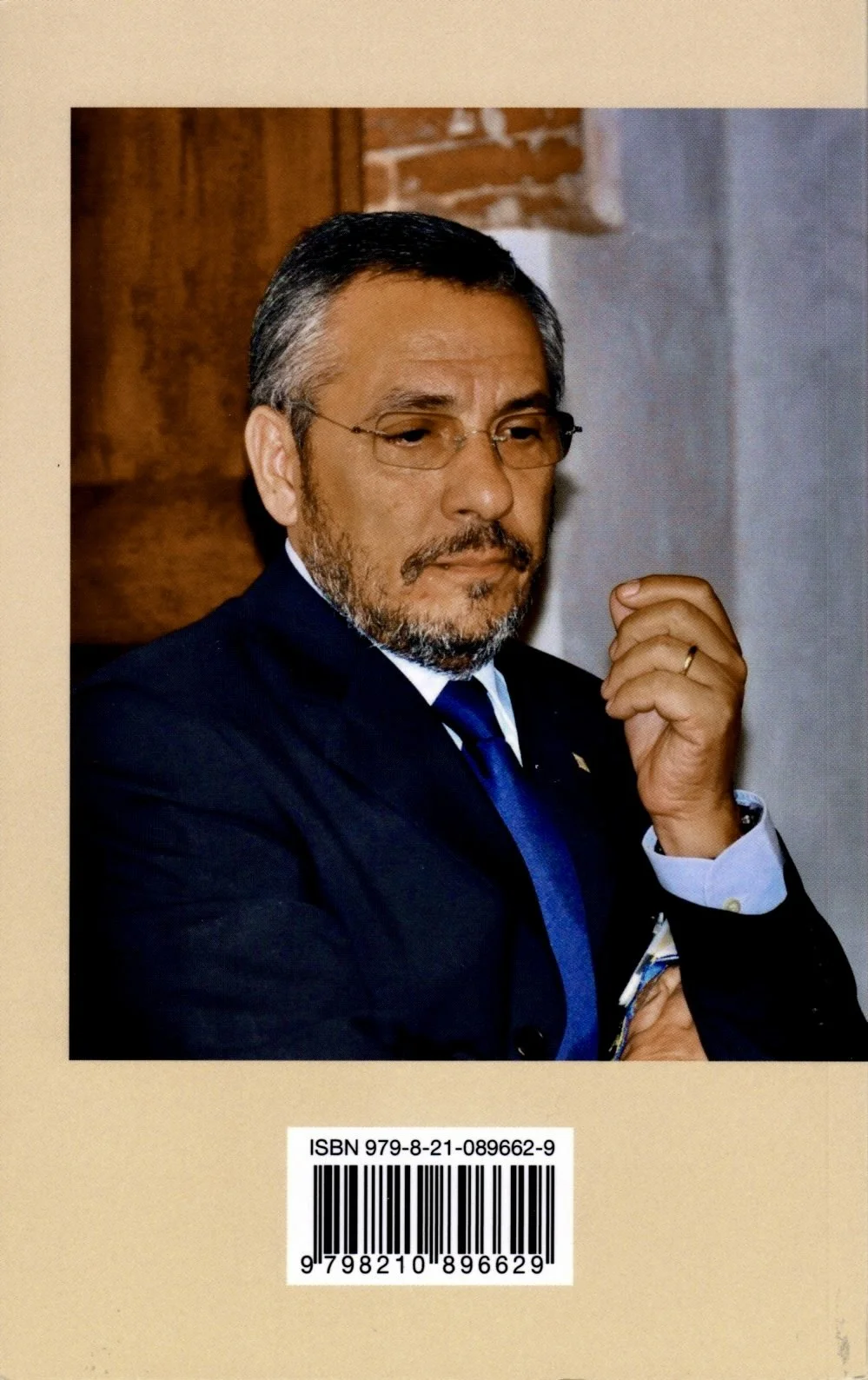 Una fotografia di un uomo con capelli e barba grigia, indossa occhiali, giacca scura e cravatta blu, sembra pensieroso, con una mano vicino al viso e una noce sul dito.